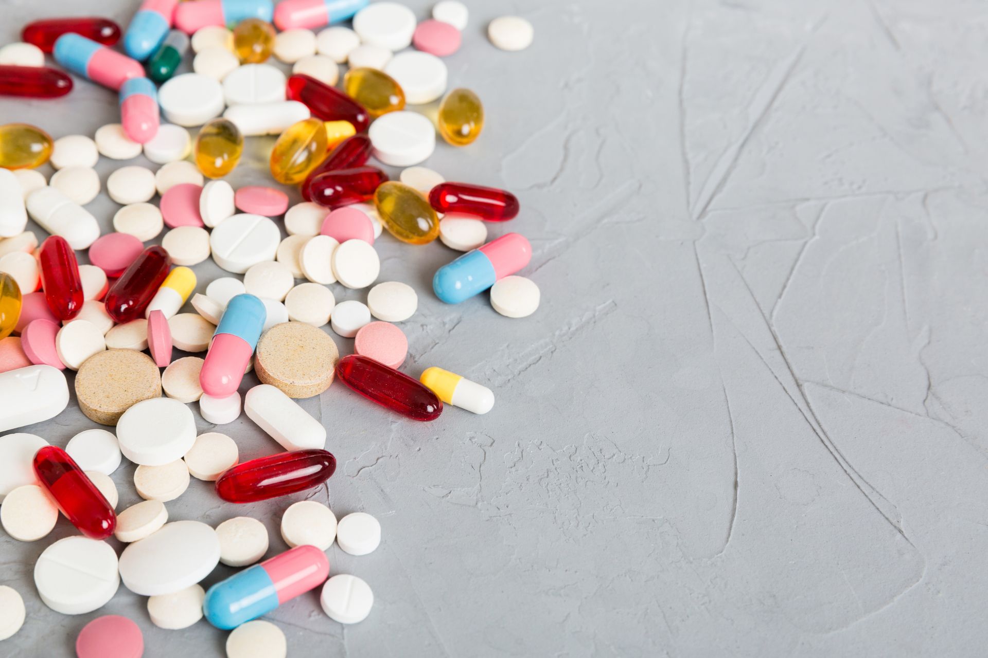 Pills and capsules scattered on a gray surface, various colors and shapes.