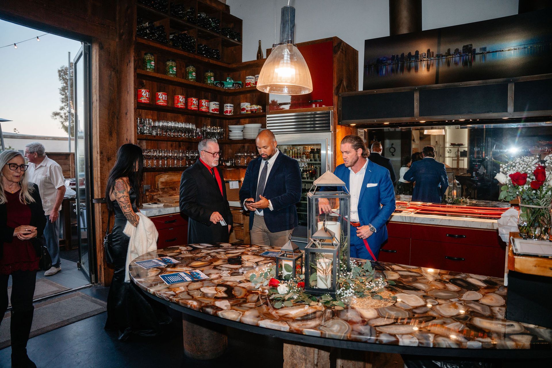 People gathered in a warmly lit restaurant. Some are standing around a table with decorative lights, and others are at the door.