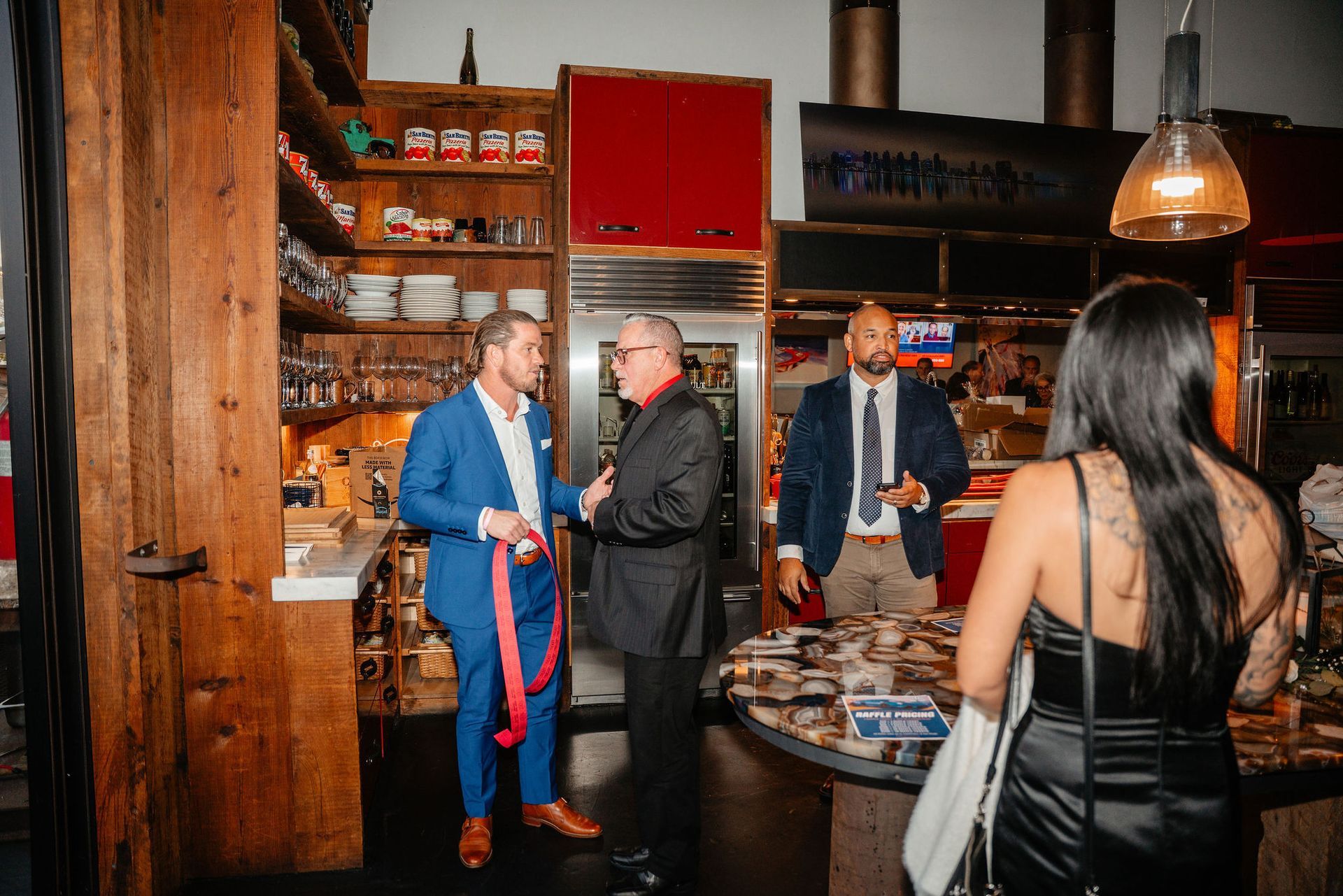 People talking at an event; indoor setting with wooden shelves and a circular table.