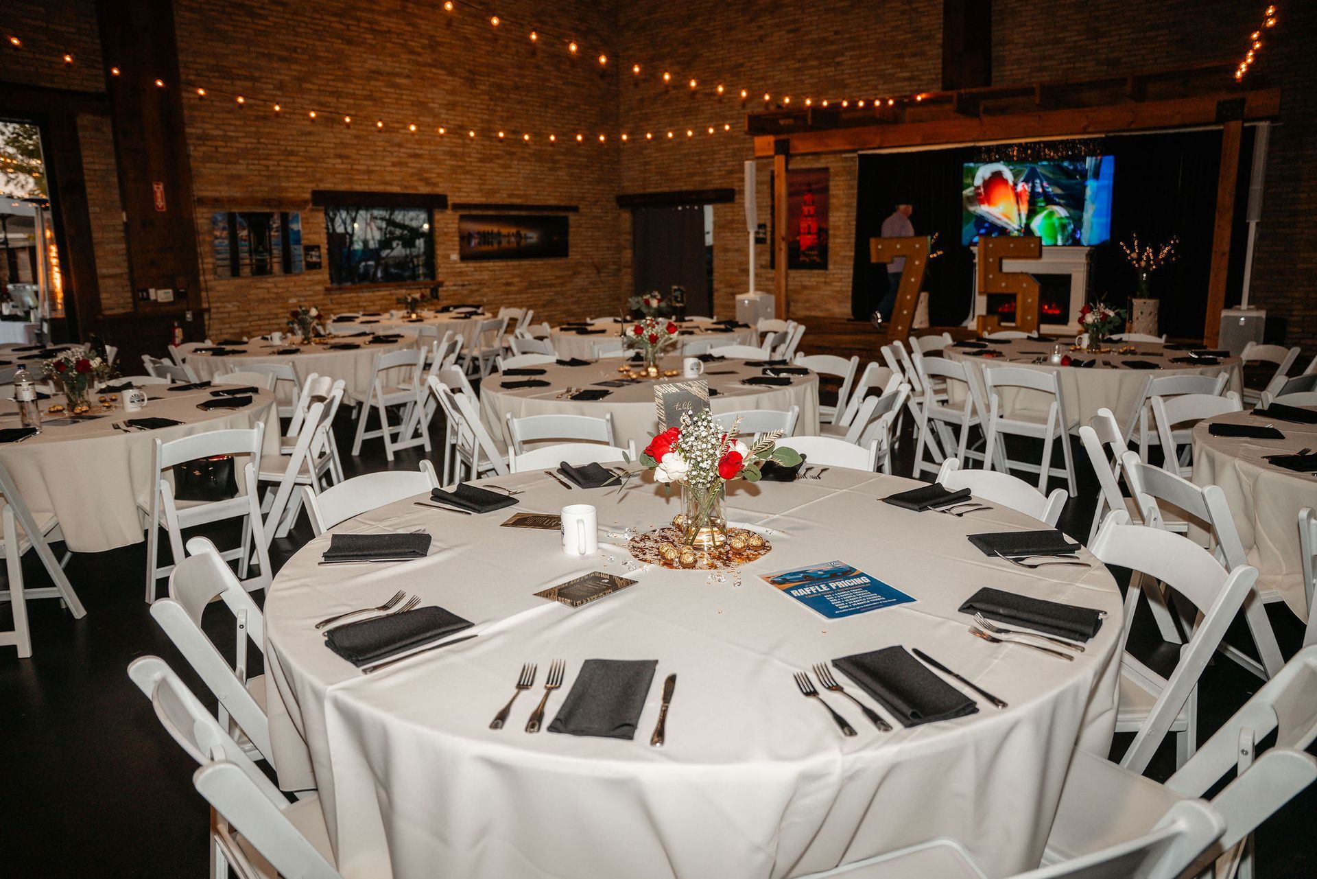 Tables set for an event with white tablecloths, chairs, and centerpieces, in a room with a stage.