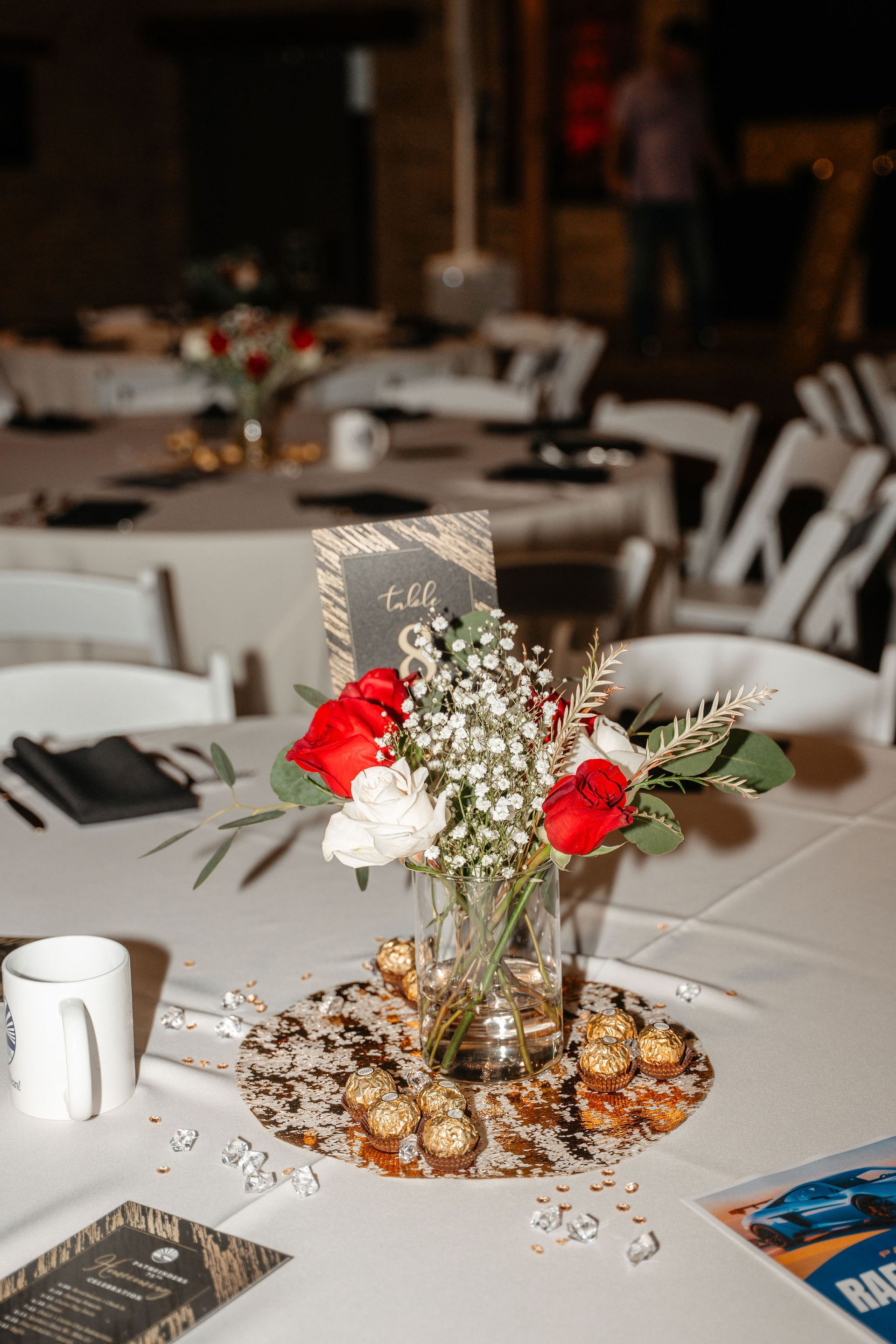 Table setting with flower arrangement, red roses, white flowers, and chocolates at an event.