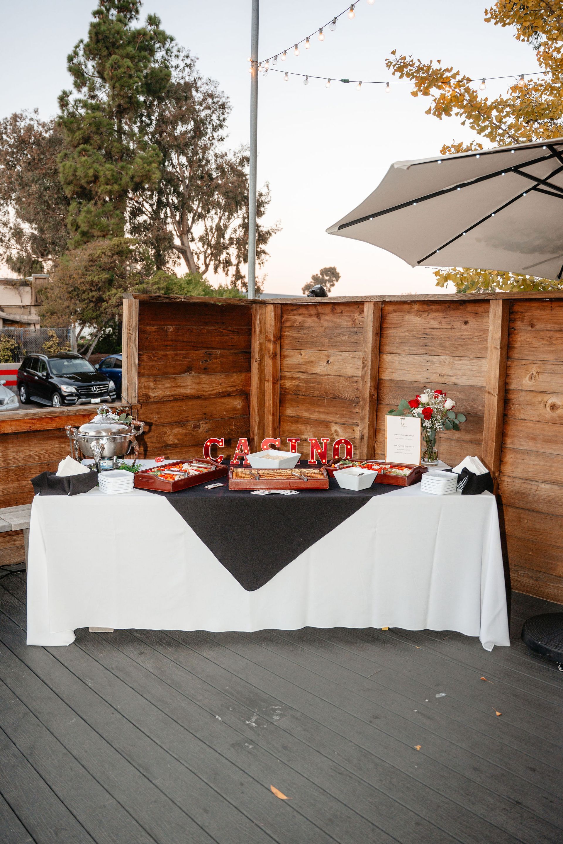 Buffet table with food on a deck, wooden backdrop, umbrella overhead, evening.