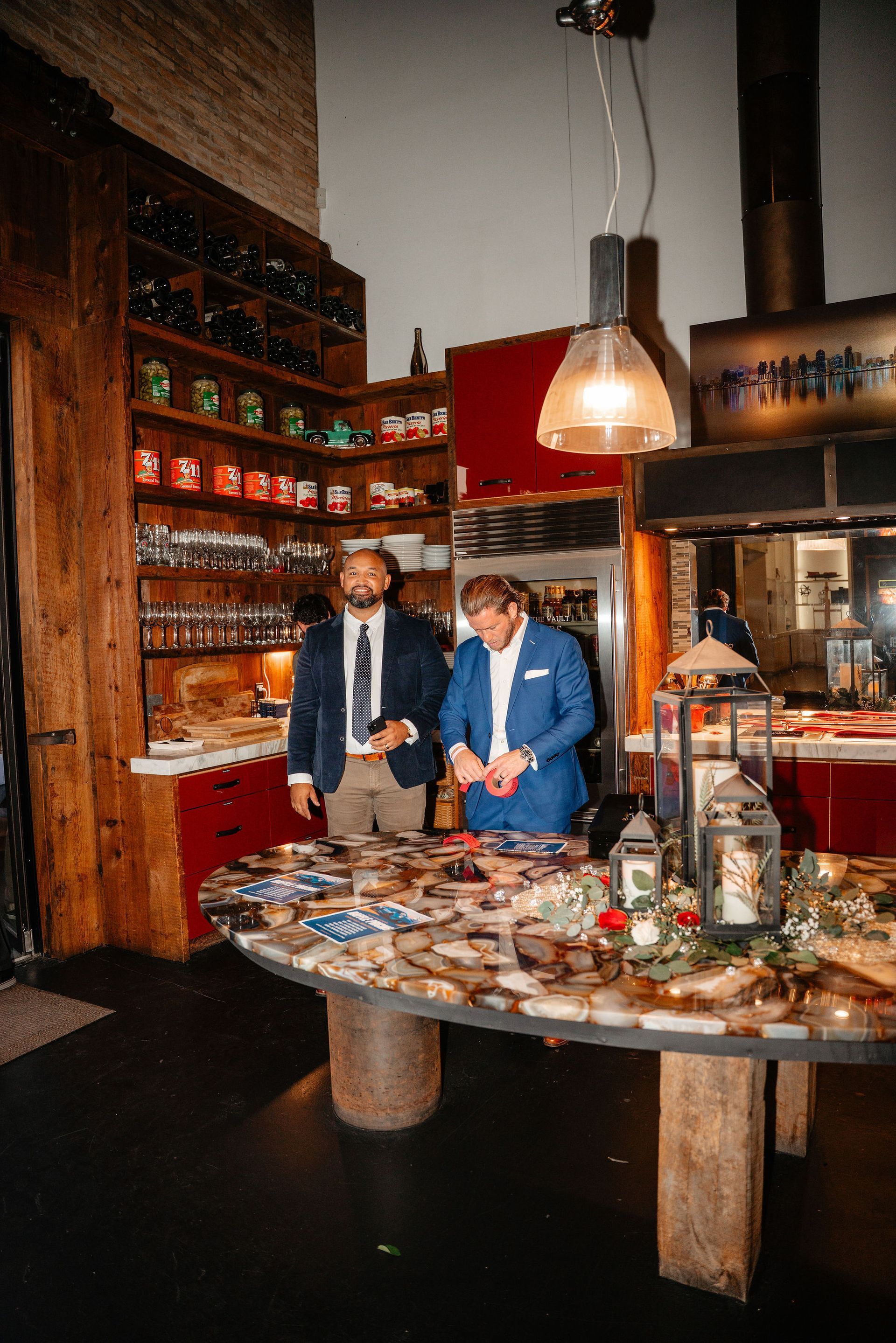 Two men standing at a table with food, shelves of items, and a red wall in a restaurant setting.