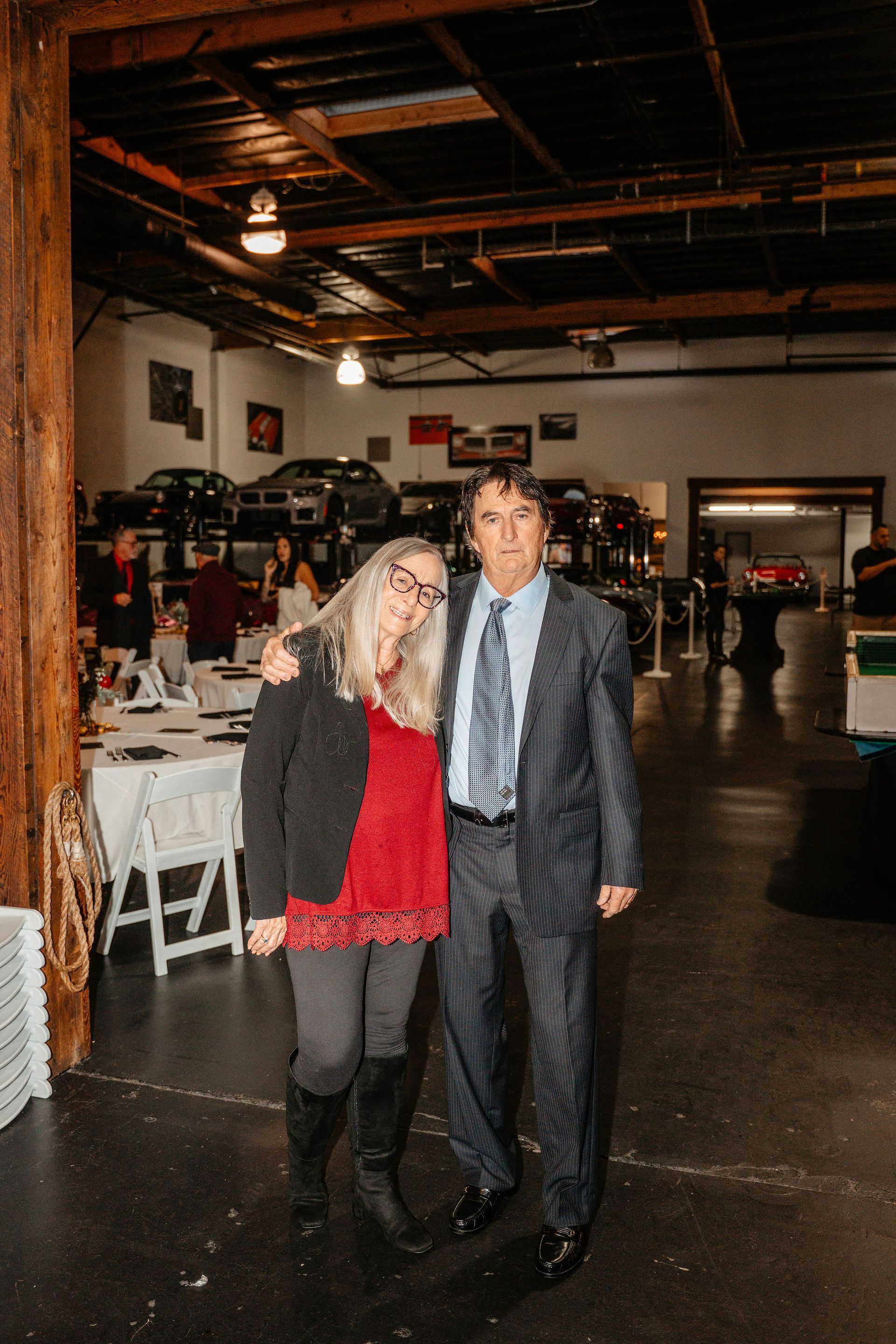 Couple posing inside a car showroom. Woman in red top and black pants. Man in suit. Tables and cars in background.