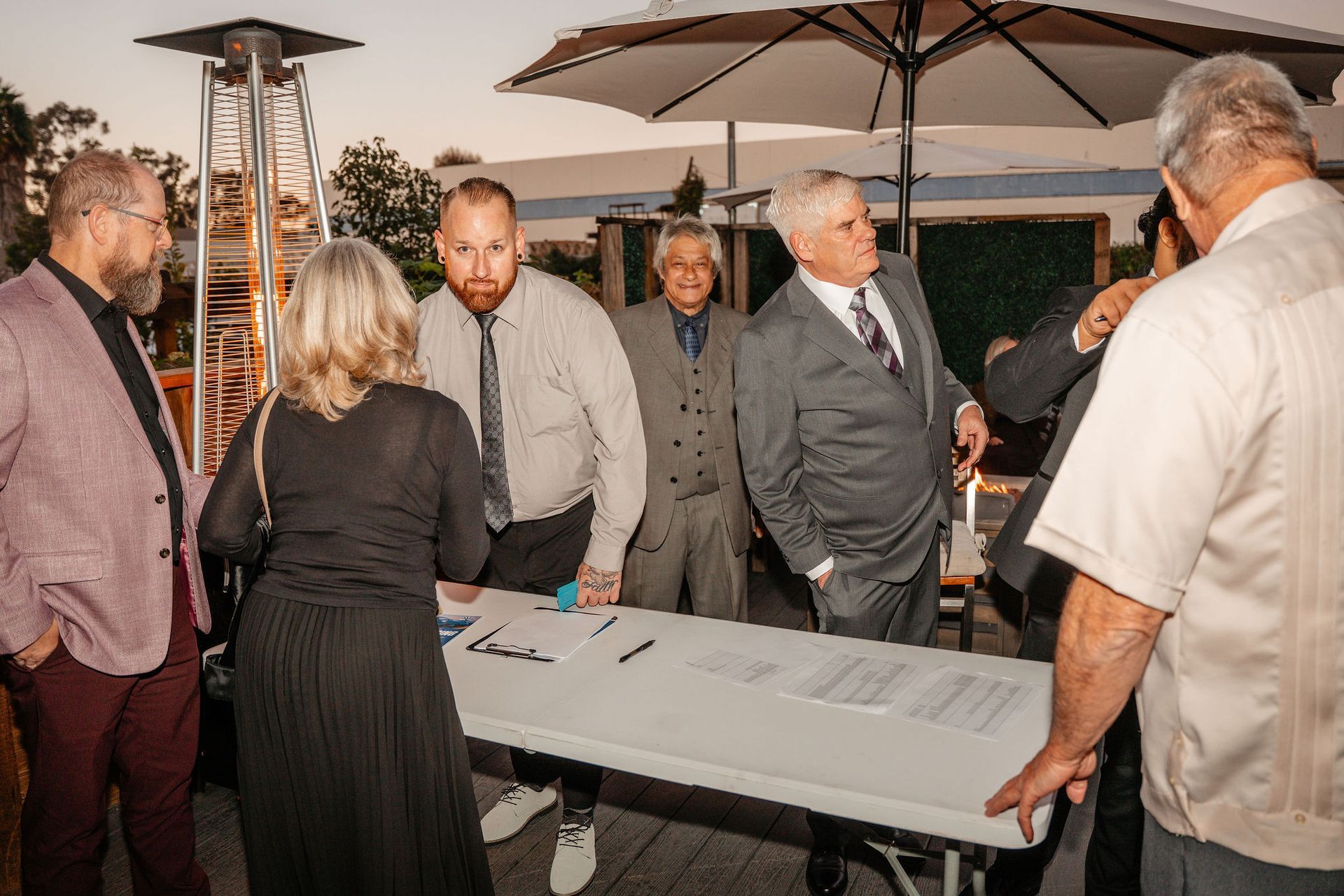 People standing at a registration table outdoors. One woman in black, men in suits, conversing.