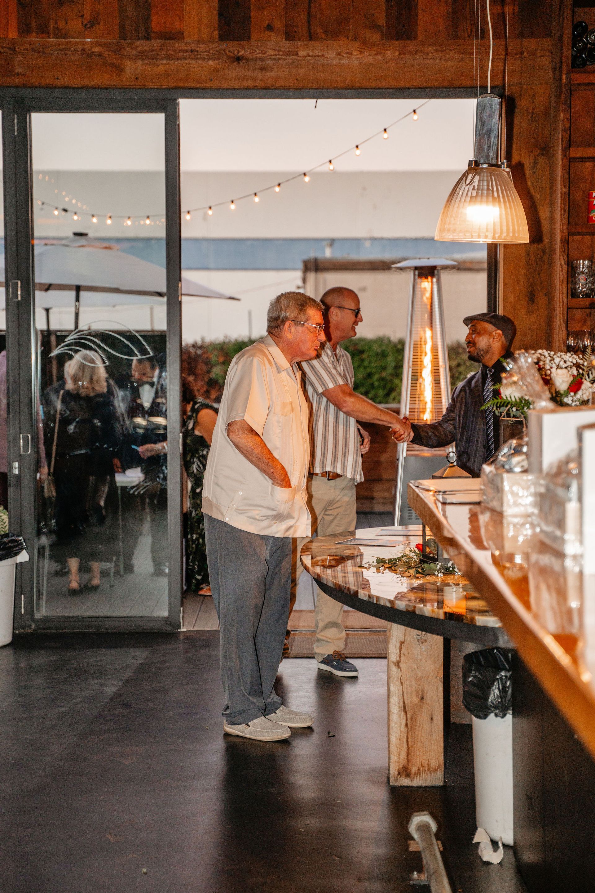 Two men at a bar, shaking hands with a bartender. An open door leads to an outdoor area.