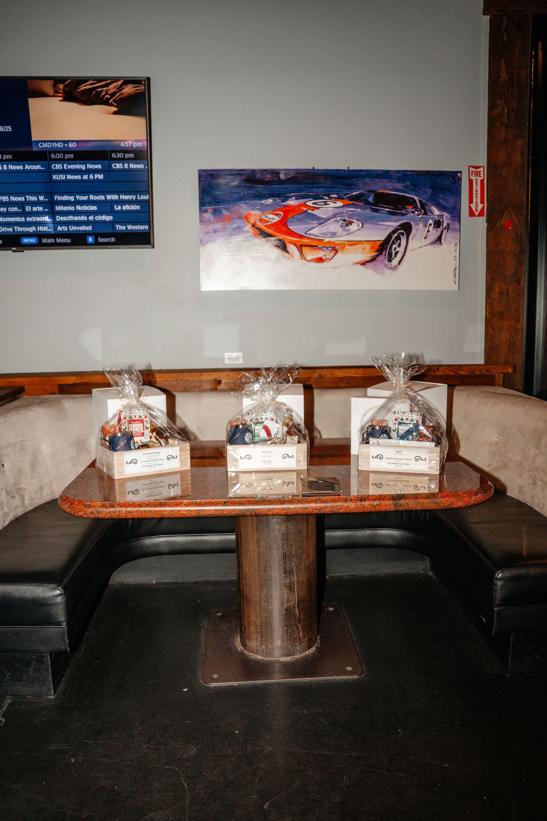 Three gift baskets on a table in a bar; car art and a TV screen on the wall.