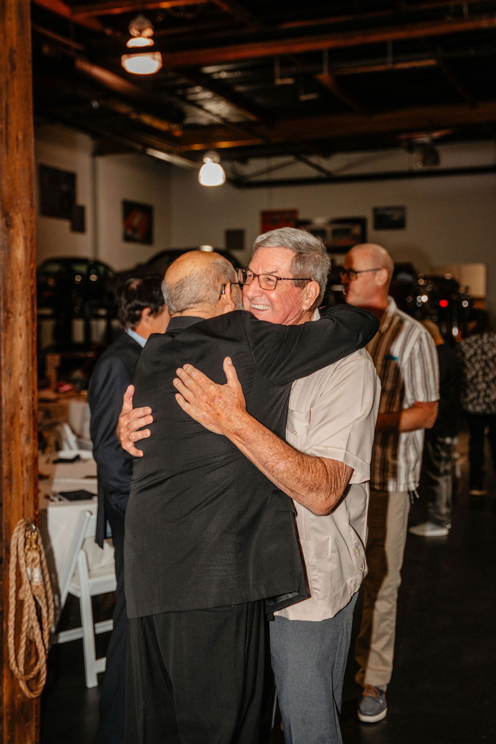 Two men embracing indoors, warm lighting. Others gather in a room with industrial details.