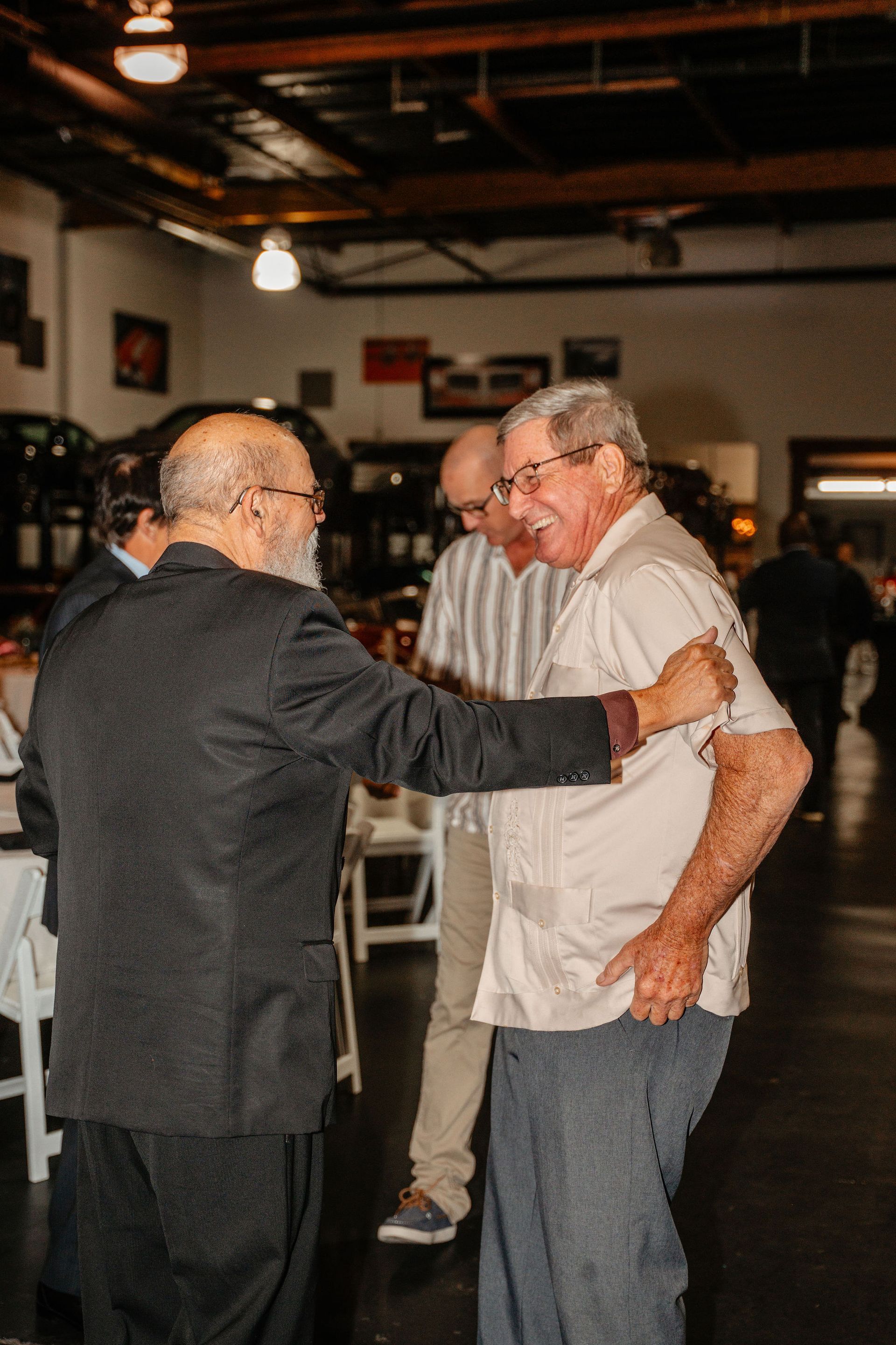 Two men greet in a room; one in a blazer, the other in a light shirt, smiling. Others stand nearby.