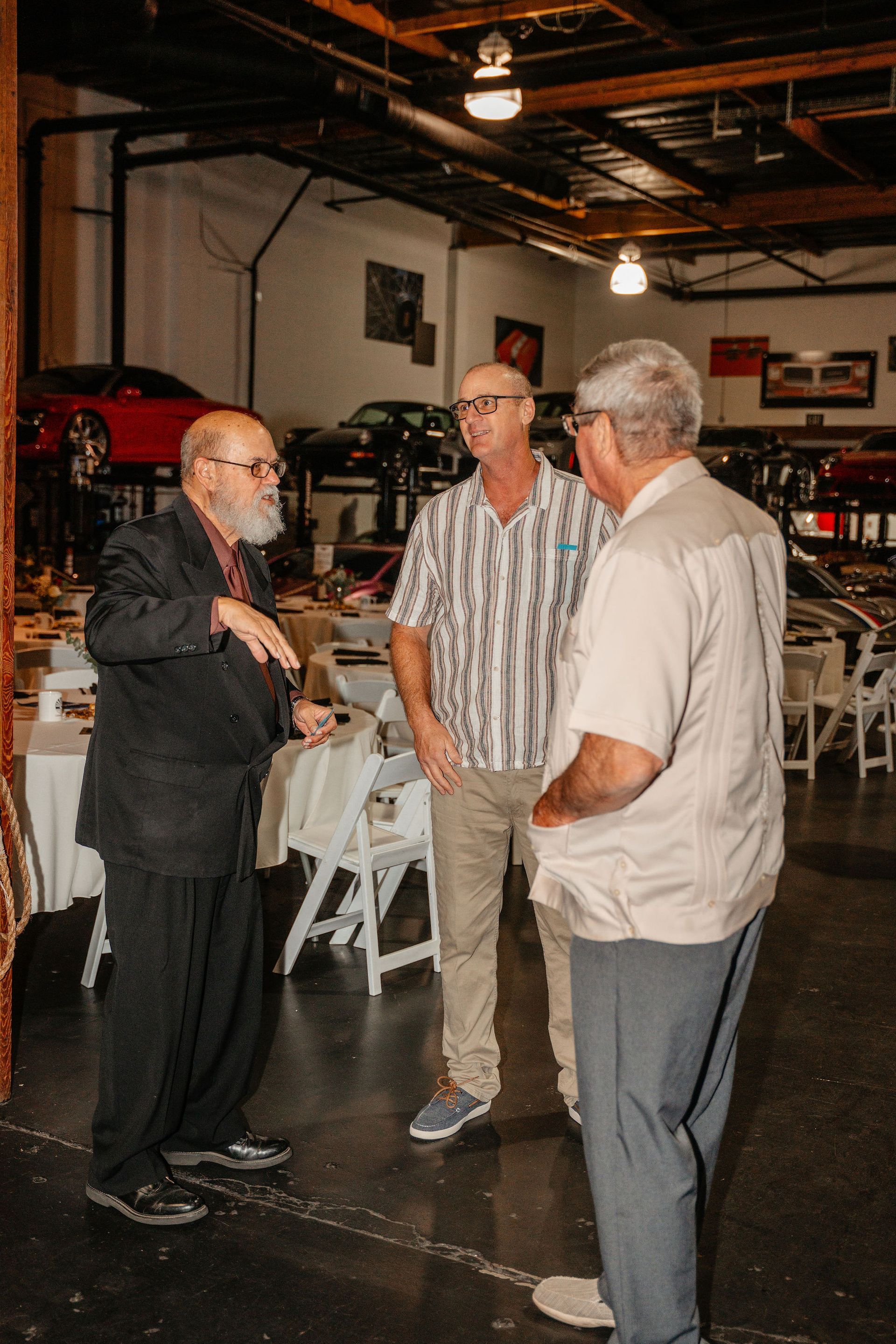 Three men talking in a garage. One gestures. Cars and tables are visible.