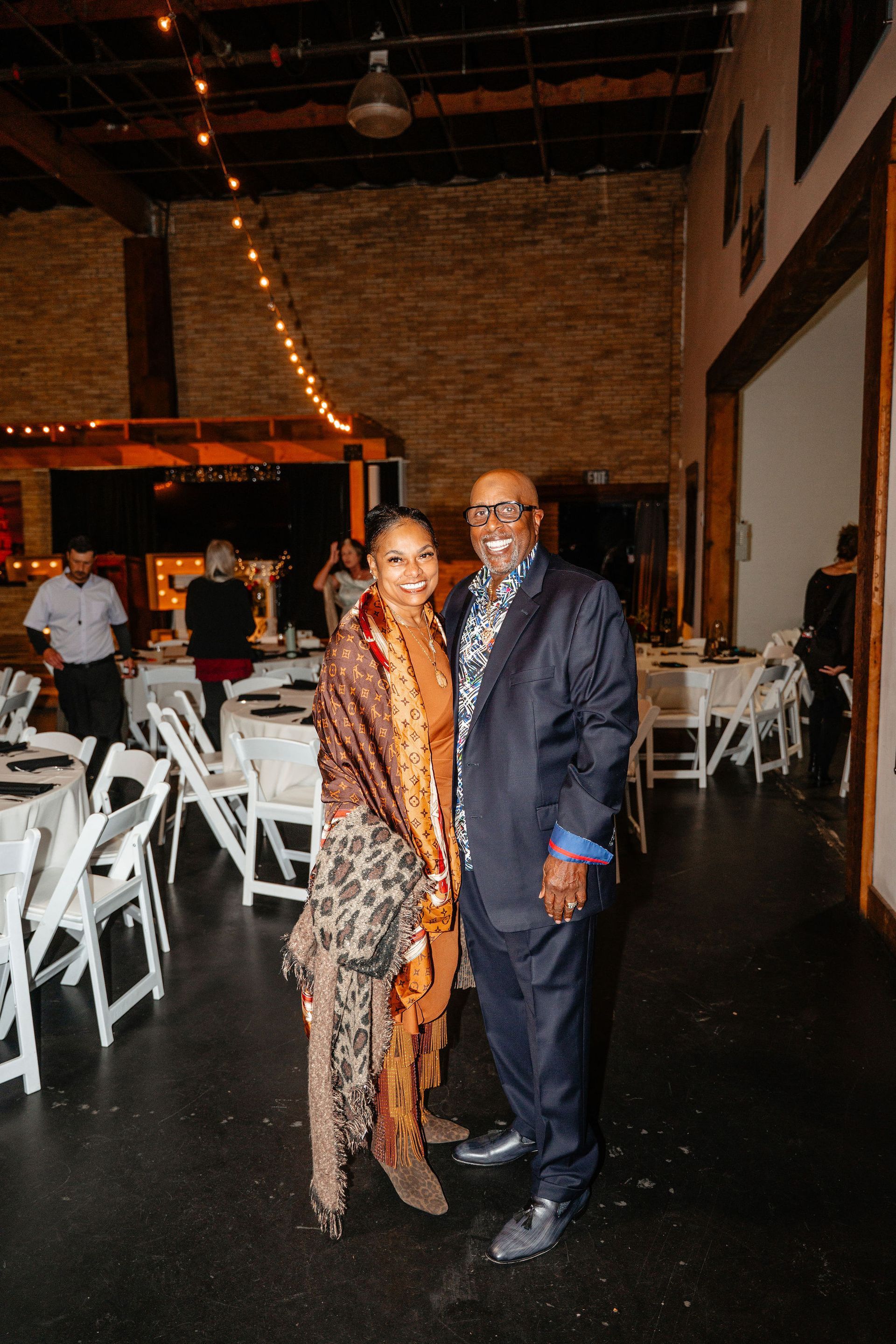 Couple posing at an event. Woman in patterned shawl, jumpsuit. Man in blazer, patterned shirt. White chairs, tables in background.