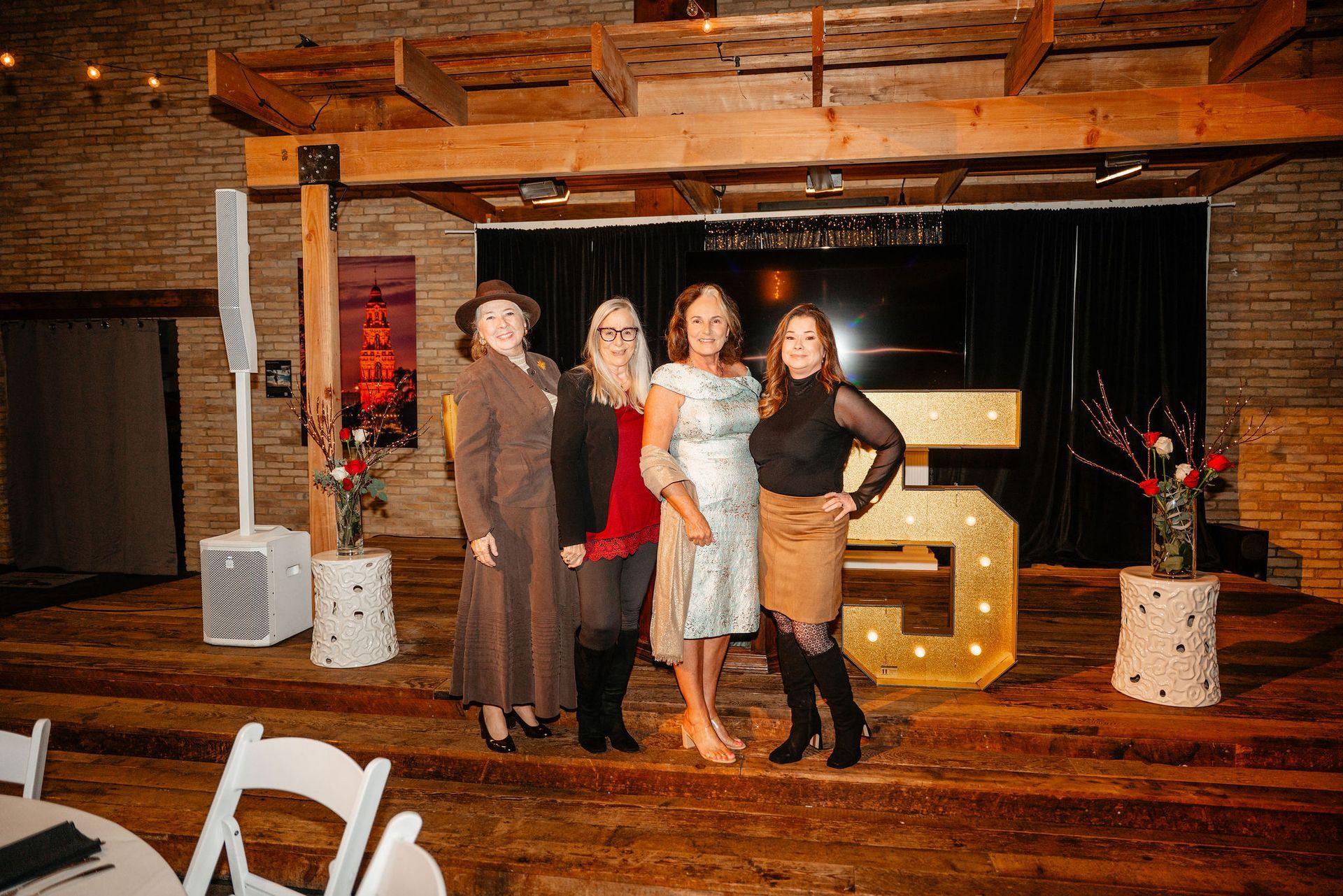 Five women pose on a stage at an event, with the letter 