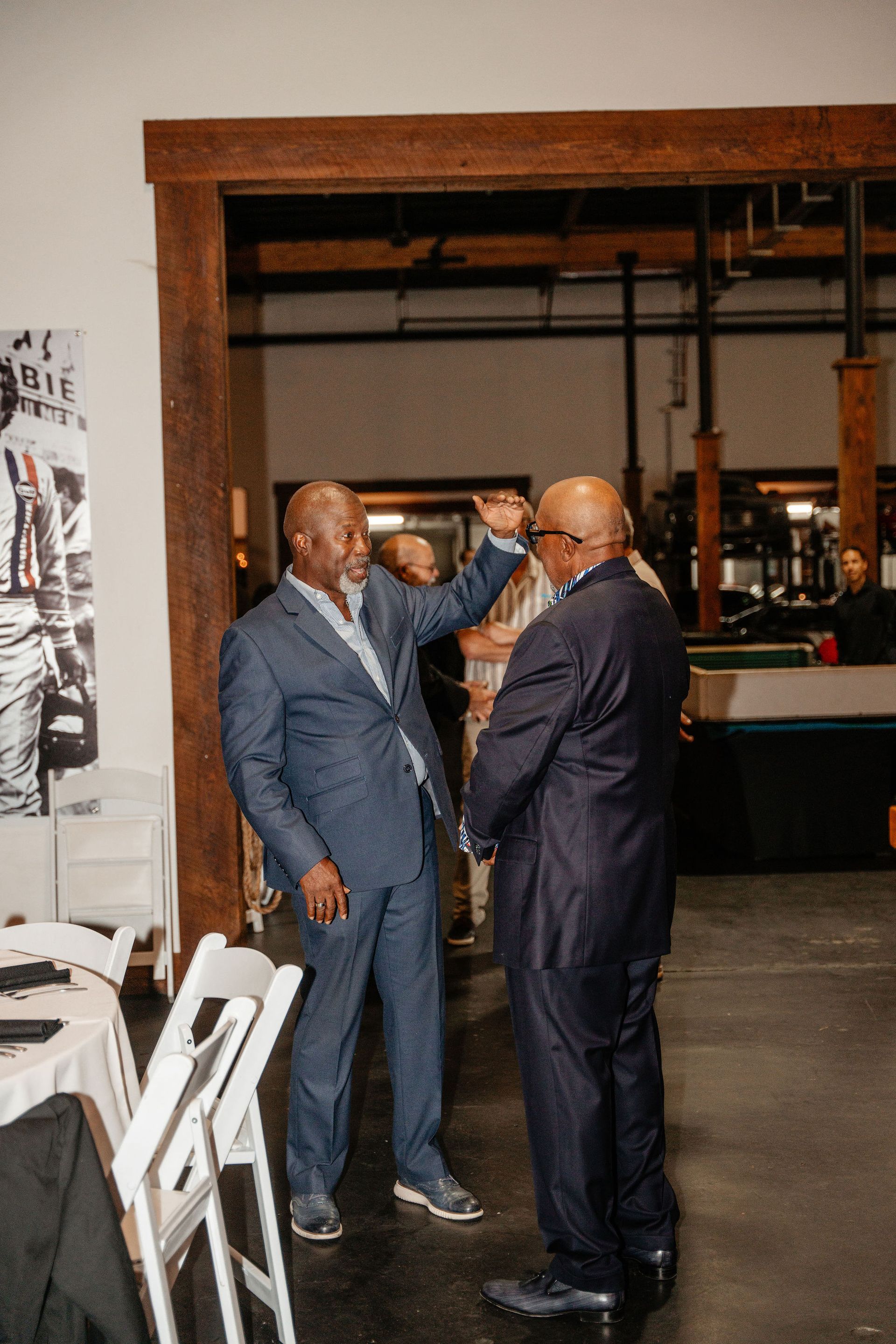 Two men in suits conversing indoors; one gestures up, the other listens.