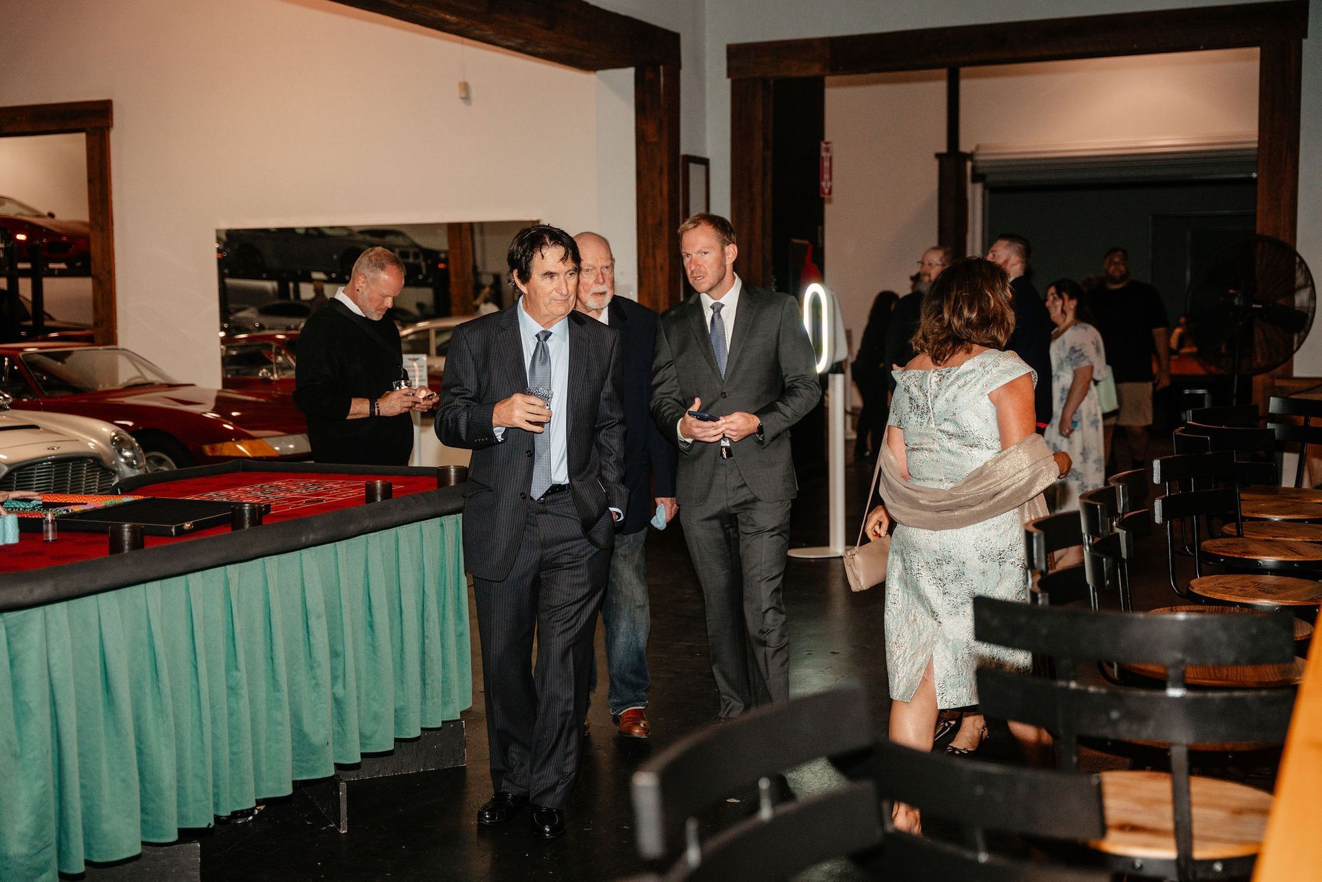 People in suits at an event, some with drinks. Red and white cars are visible. Interior setting.