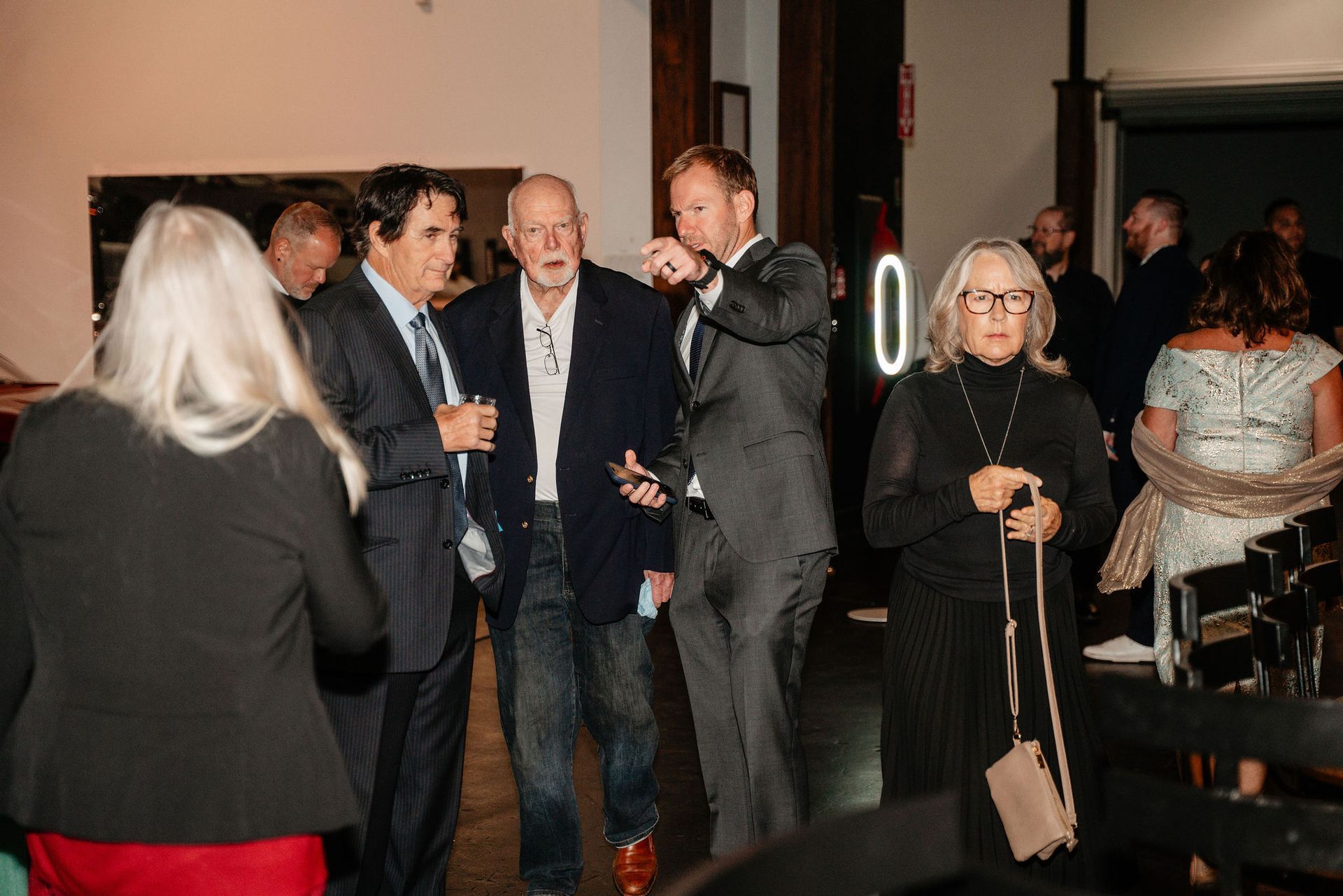 People socializing indoors; man in blazer gestures toward another man, others nearby.
