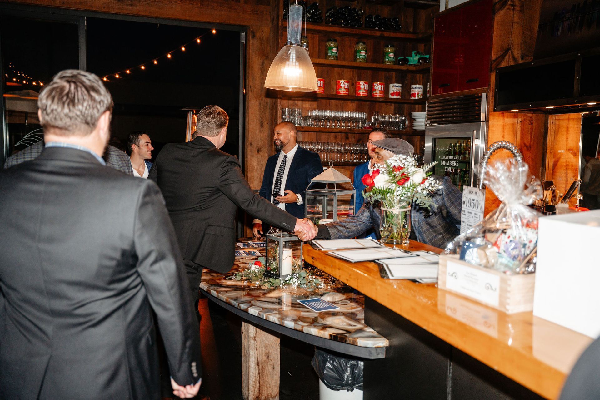 People in suits shaking hands at a bar. A man in a suit approaches the bar. Festive decor is visible.
