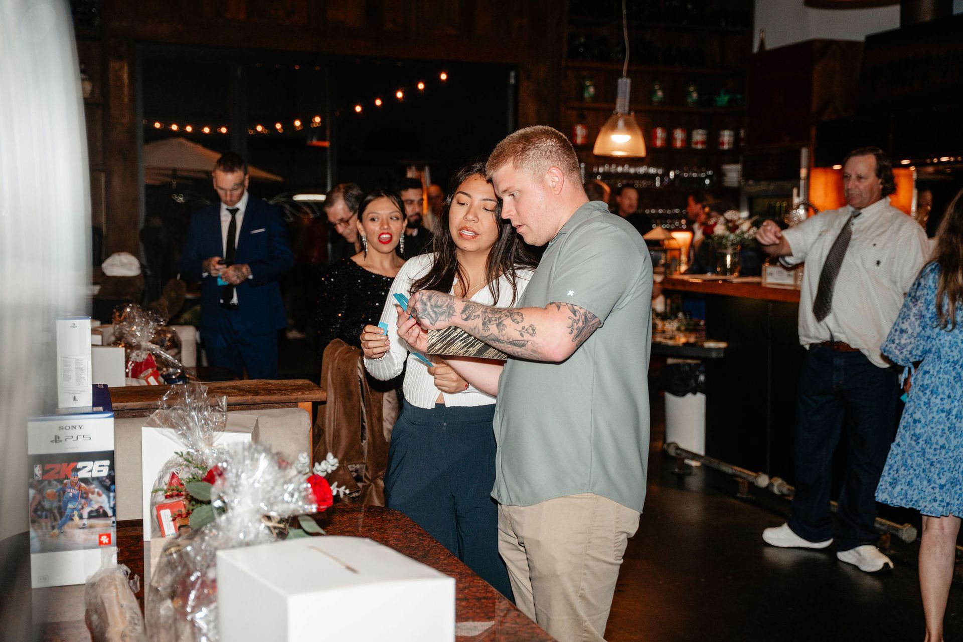 People at event, examining items on table. Man with tattoos, woman, and others look at something in his hands.