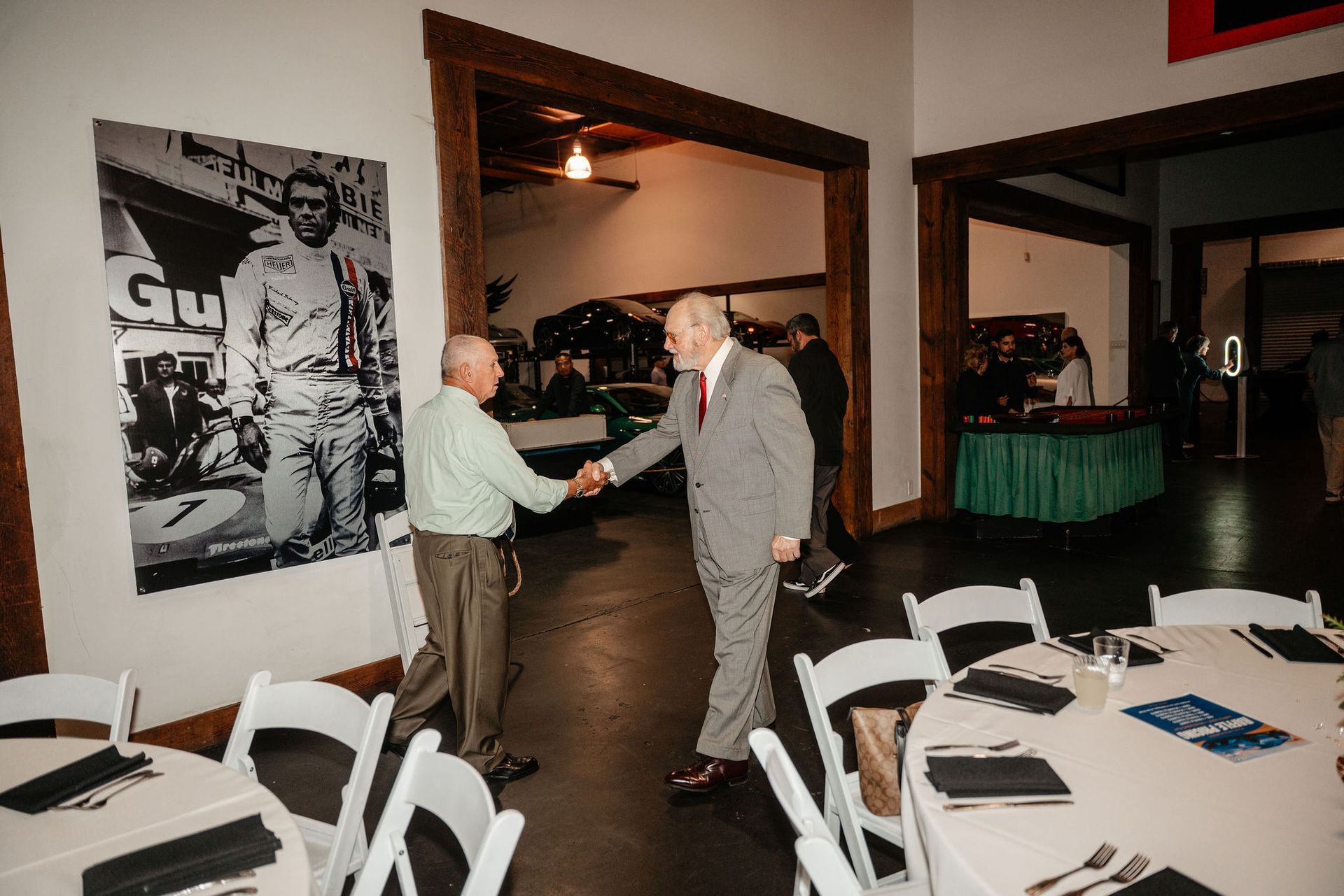 Two men shaking hands in a room with tables and a race car poster.
