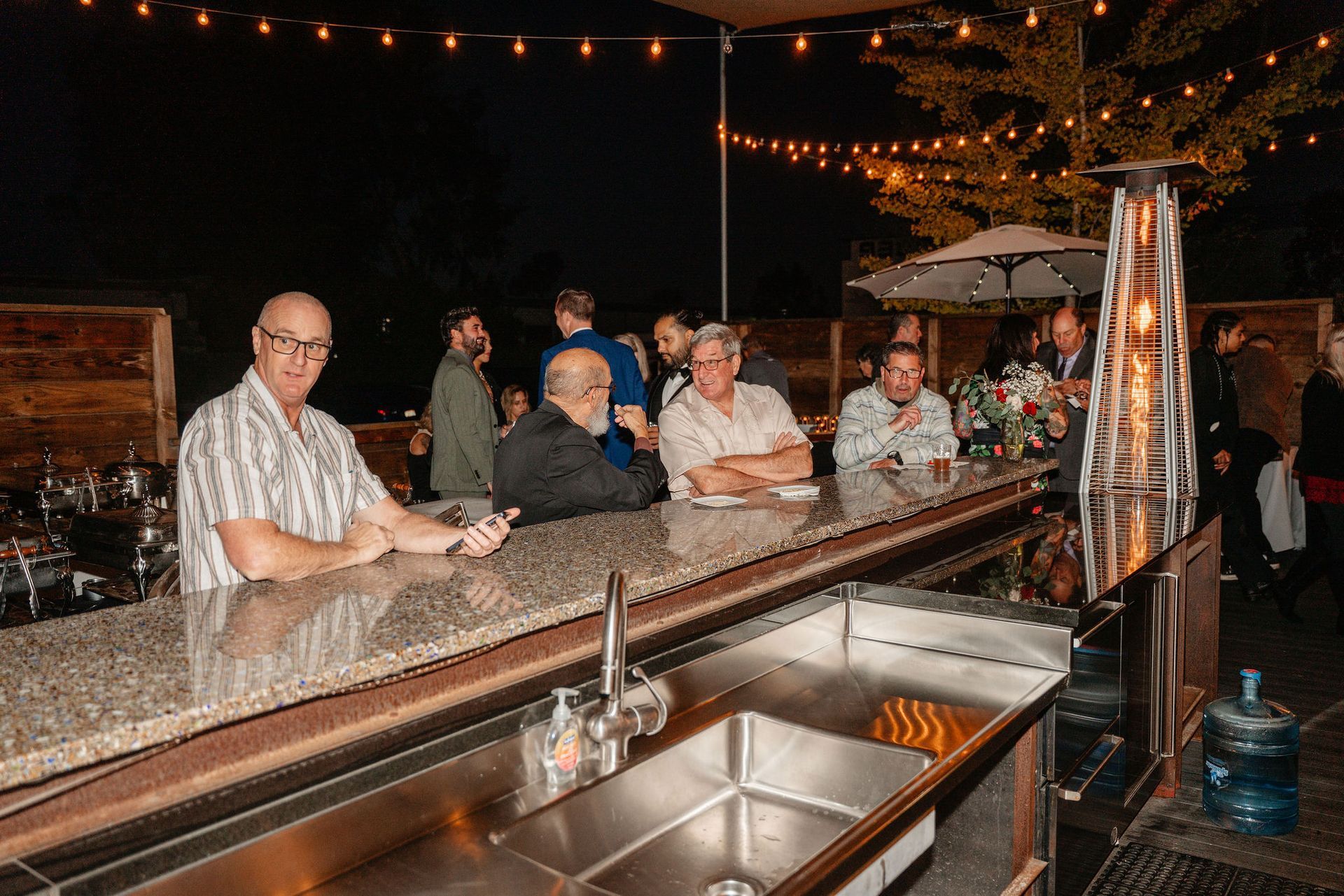 People at an outdoor bar at night. Strings of lights overhead. A man at the counter looks at the camera.