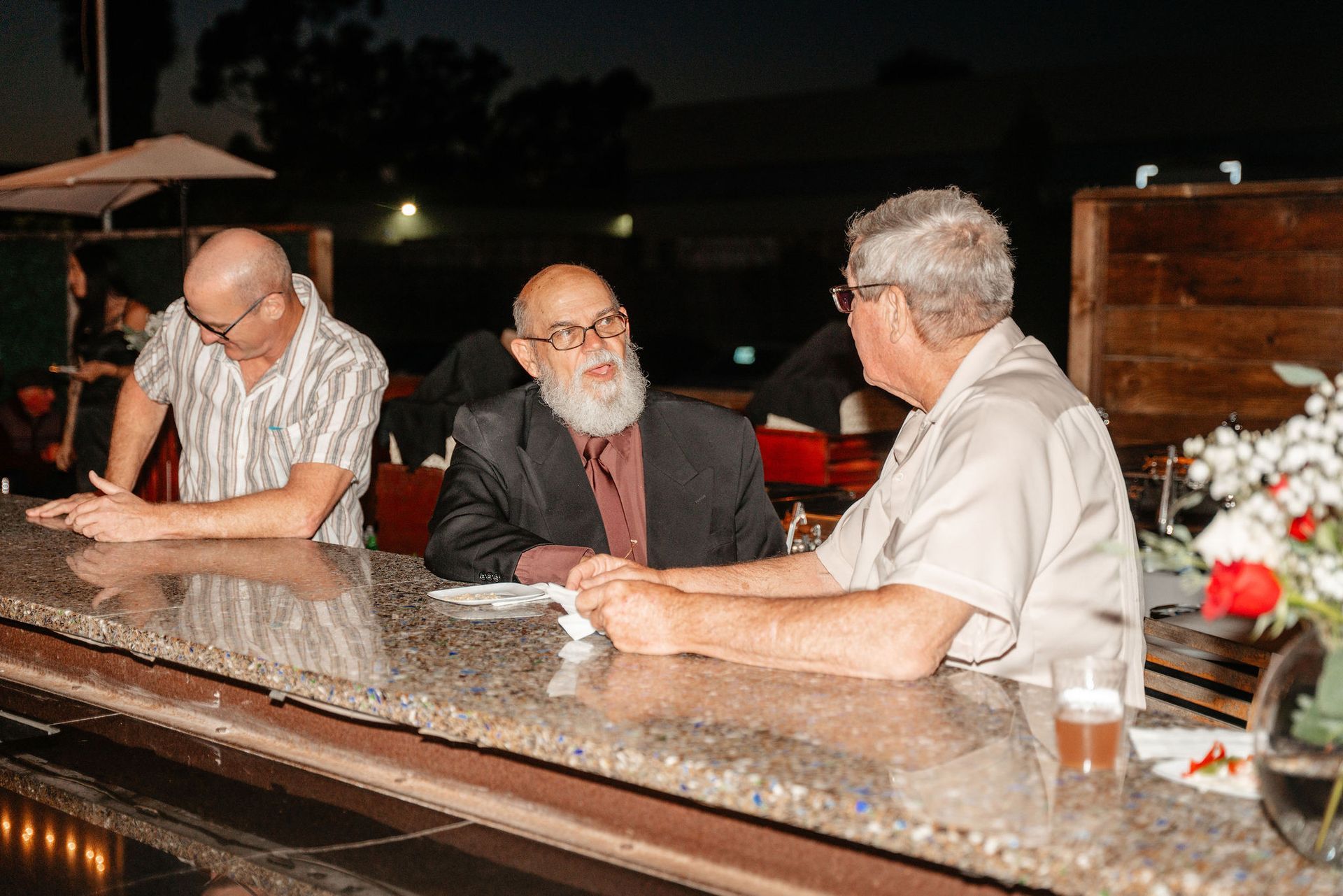Three men conversing at a bar. One with a long beard and suit, two in casual shirts. Night setting.