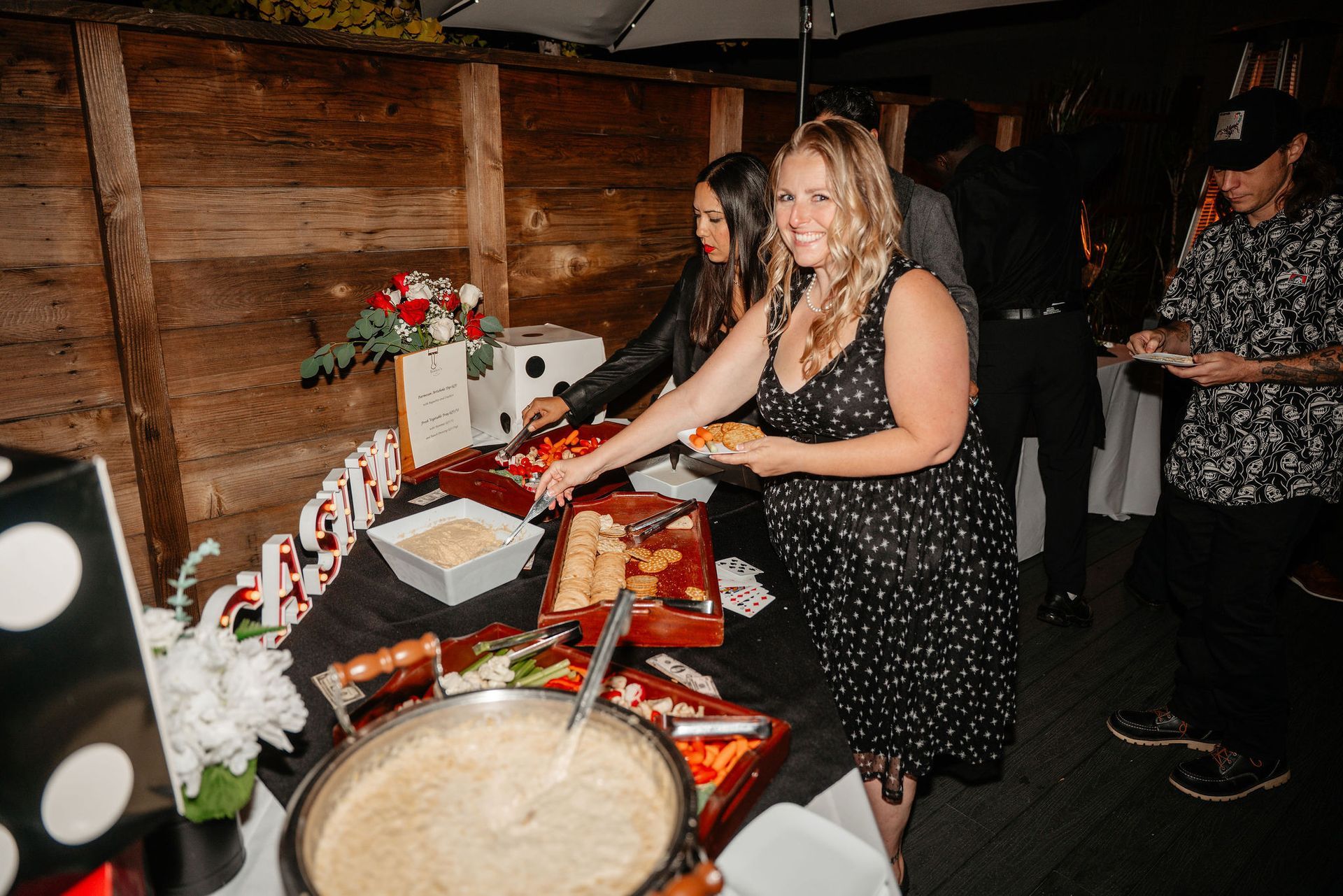 Woman in black dress at a food buffet, reaching for food. Others are present at a table with various dishes.