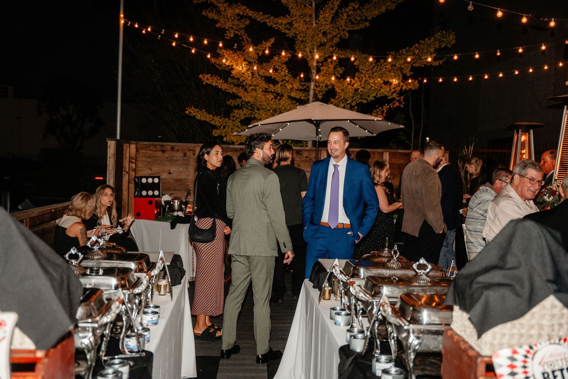 Outdoor evening event with guests, buffet tables, and string lights.