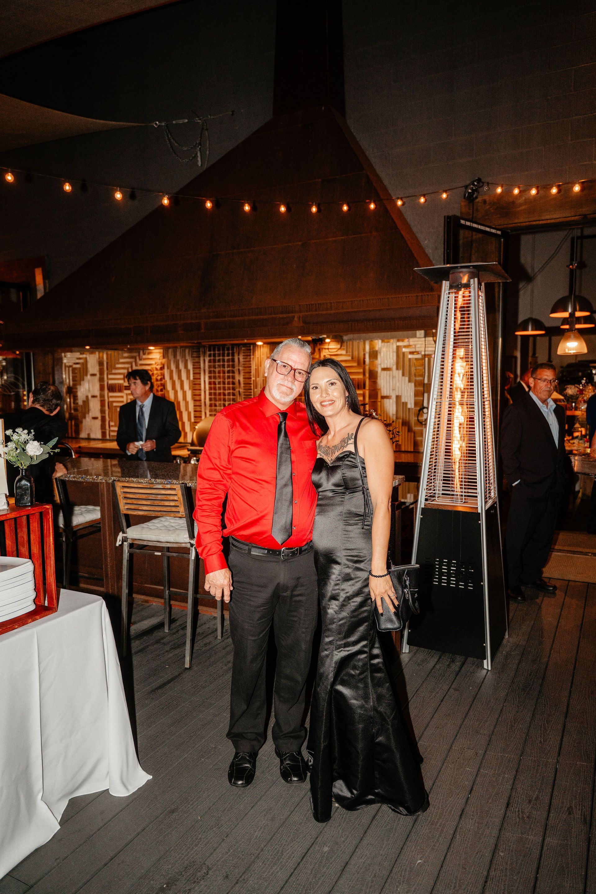 Couple posing at a party; man in red shirt and tie, woman in black dress, near a heater.
