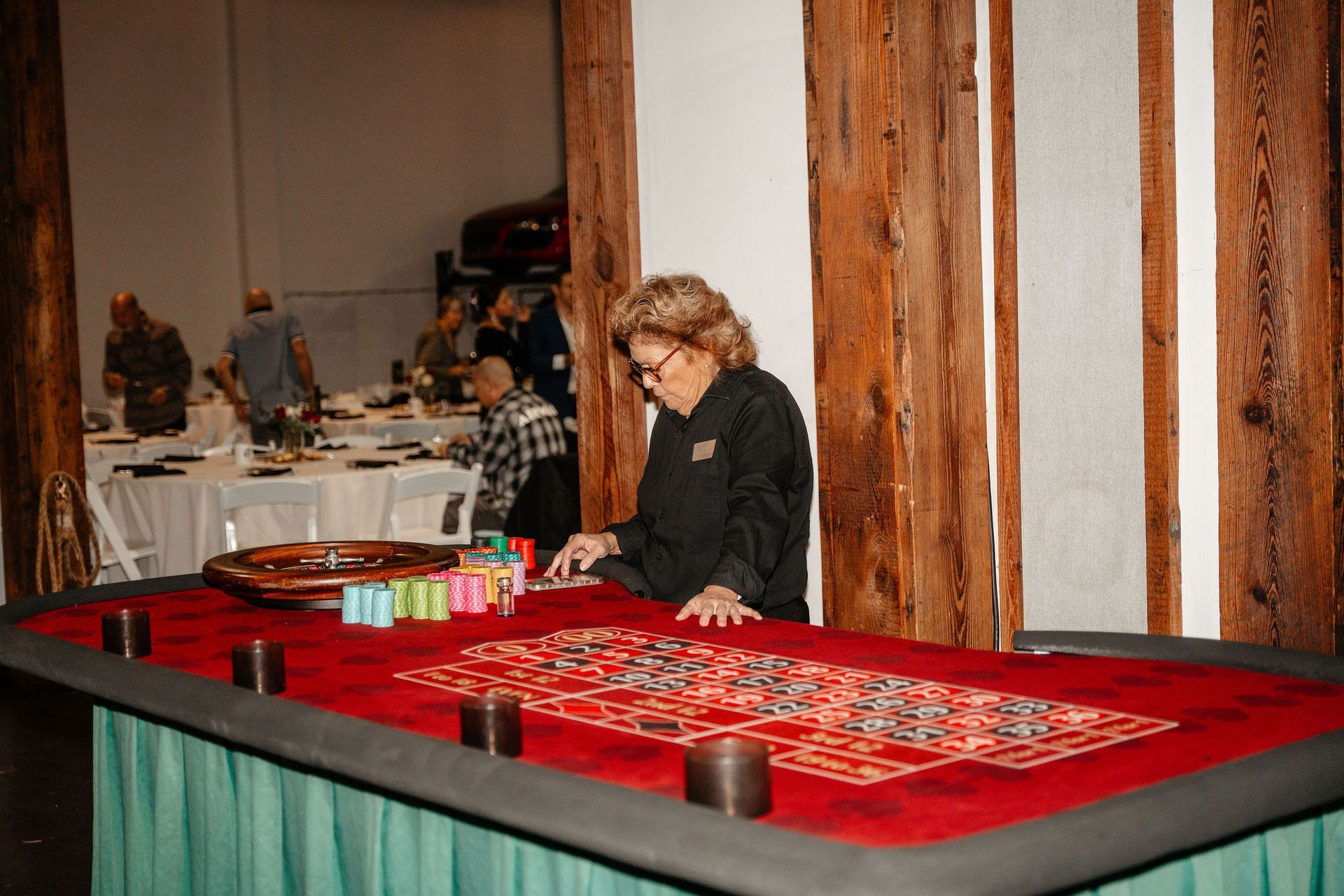Woman at a roulette table in a dimly lit room, with others in the background near a table.
