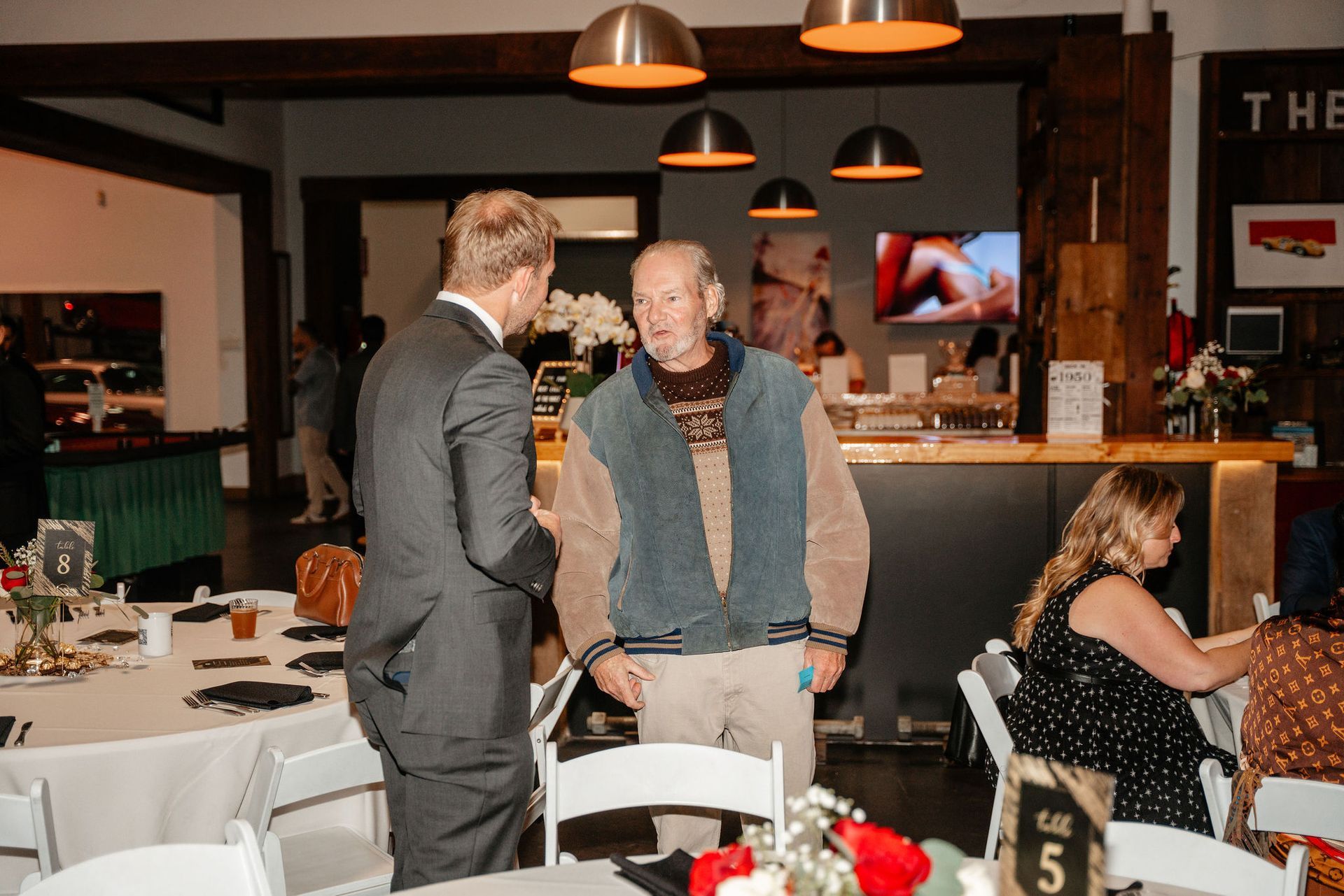 Man in suit talks to another man wearing a vest at a gathering with tables and bar in the background.