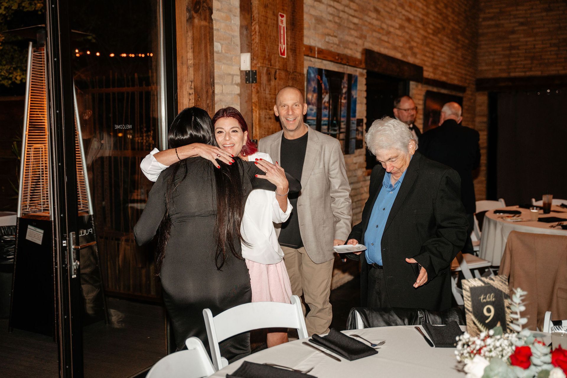People embrace at a formal event in a room with brick walls, tables, and decorations.