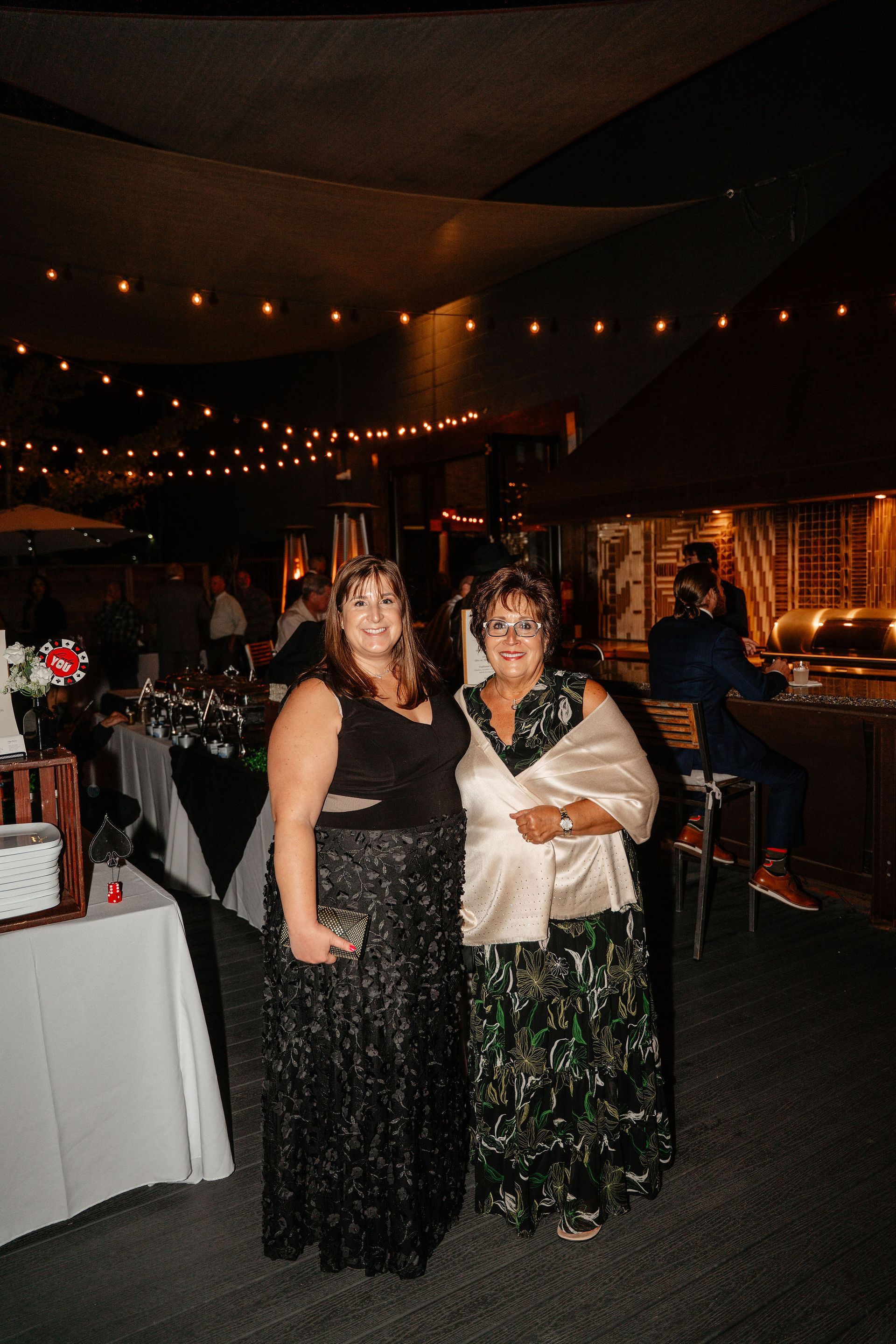 Two women at an outdoor event, one in a black sequin dress, the other in a patterned gown and shawl, under string lights.