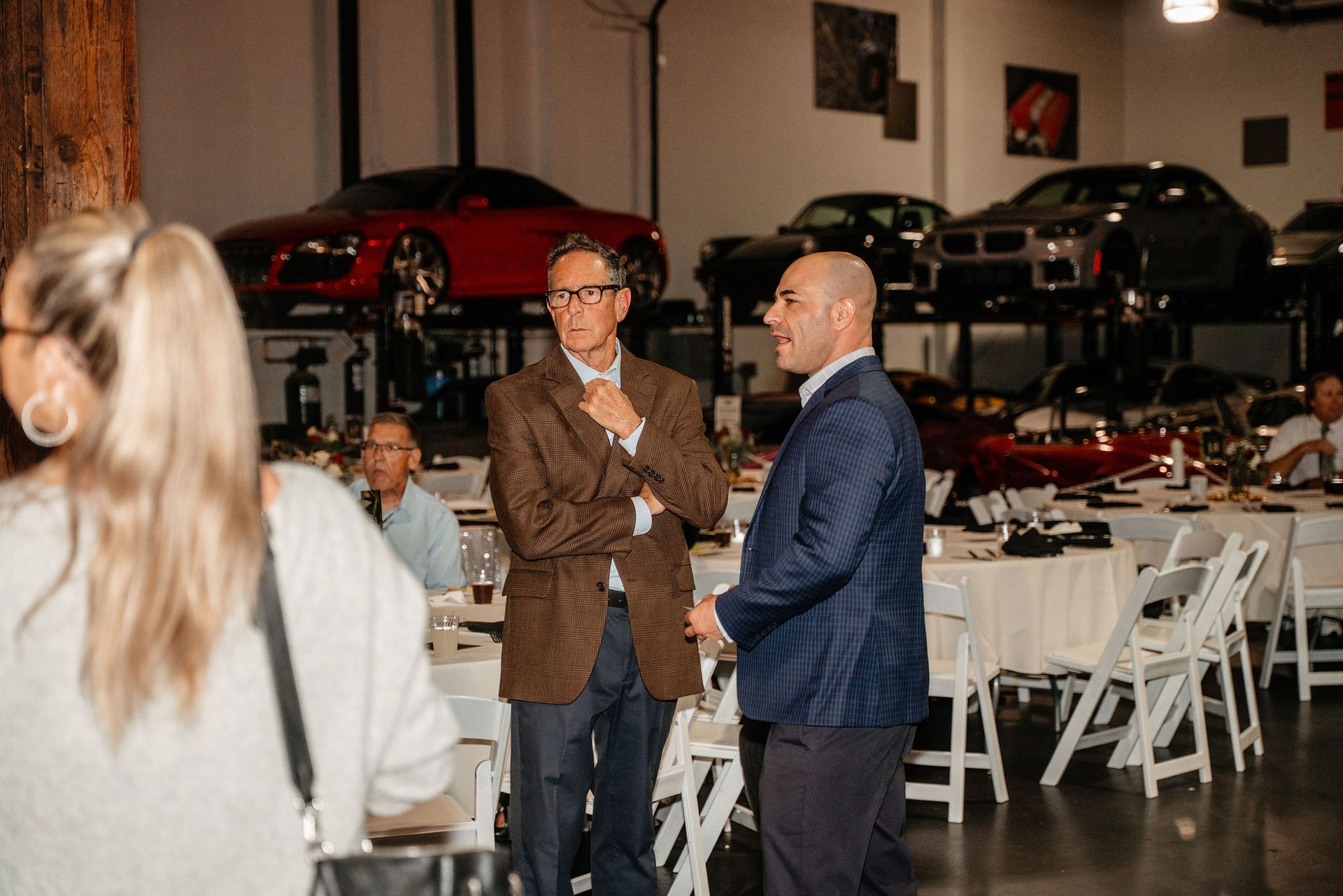 Two men in suits converse at a dinner event in a garage with cars on lifts.