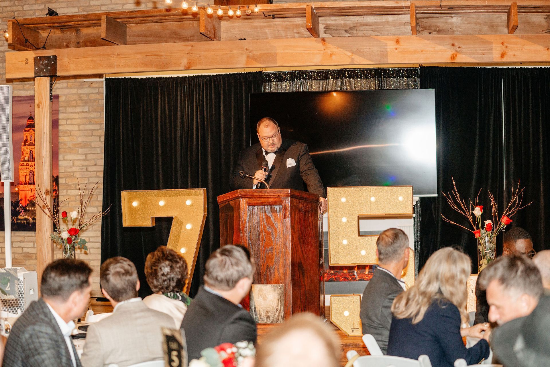 Man speaking at a podium during an event; guests sit at tables. Gold 
