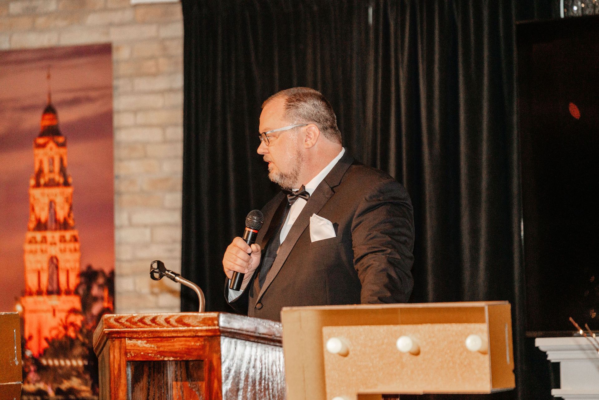 Man in a tuxedo speaking at a podium, holding a microphone. Brick wall backdrop, lit with a colorful tower image.