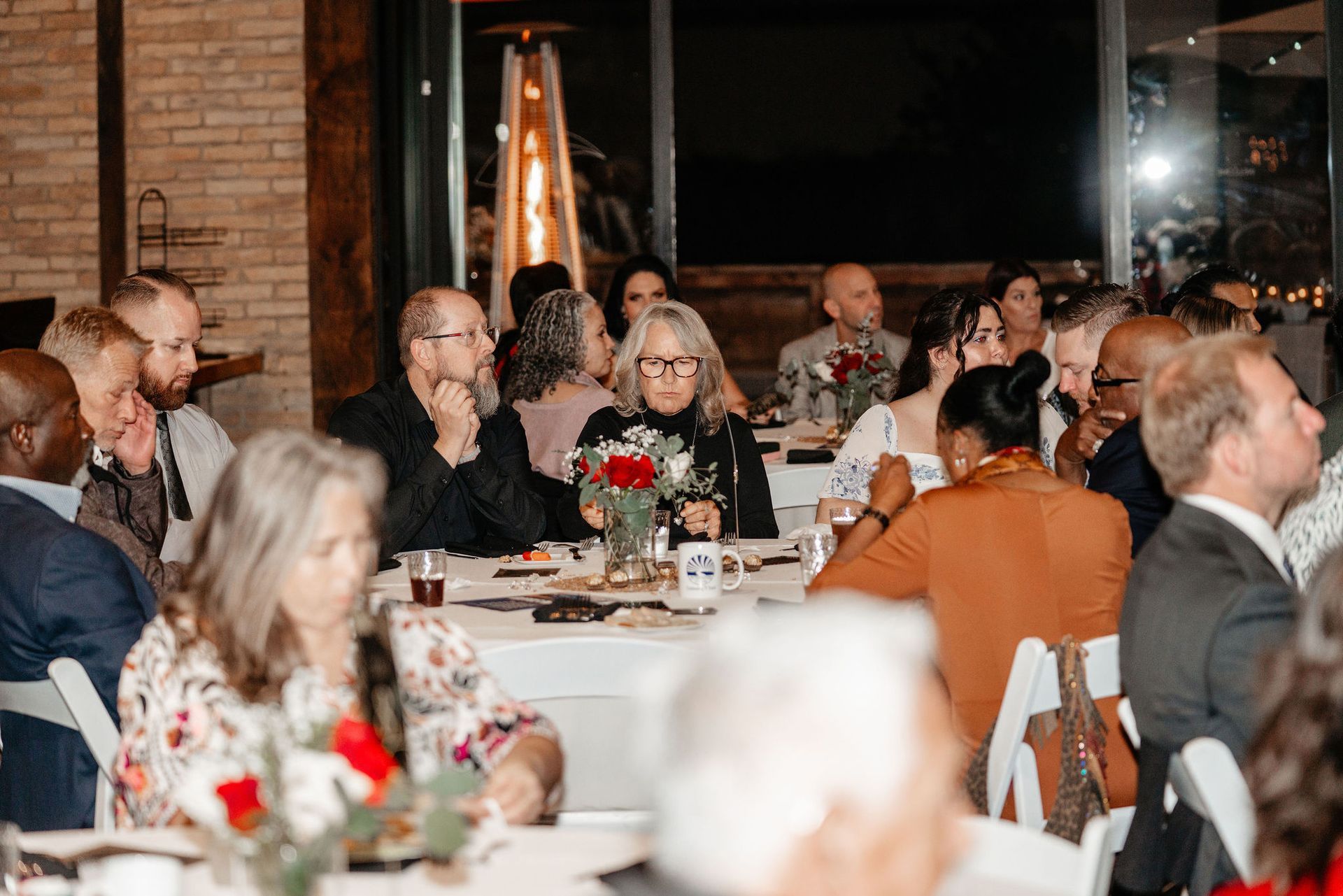 People seated at tables during an event, some looking at something unseen, with warm lighting.