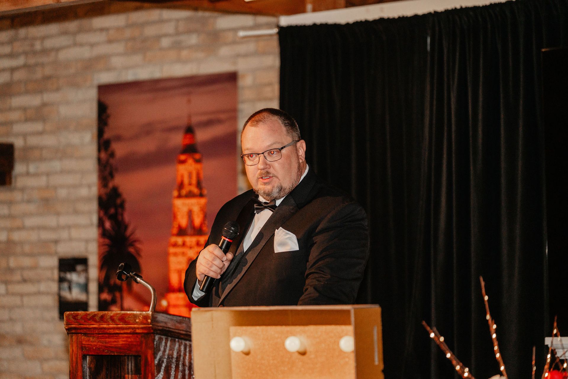 Man in tuxedo speaking at podium, holding microphone. Background has brick wall, draped curtain, and a cityscape painting.