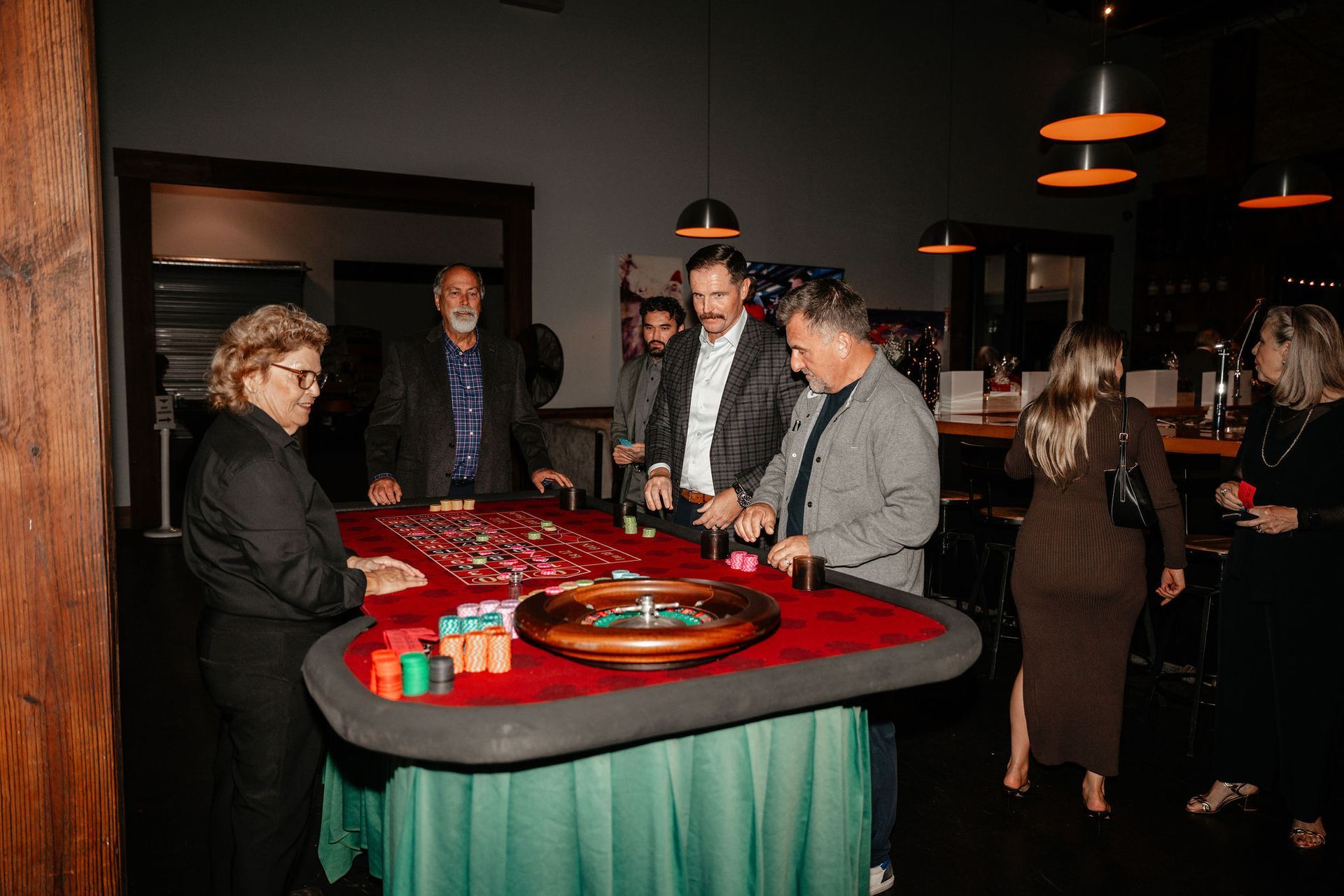 People gathered around a roulette table with casino chips. Dark room, bar in background.