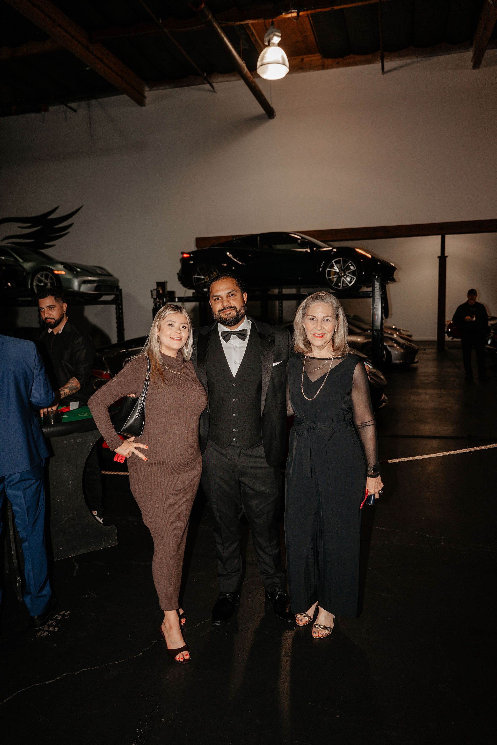 Three people pose in a garage with cars. Man in tuxedo, women in dress and jumpsuit.