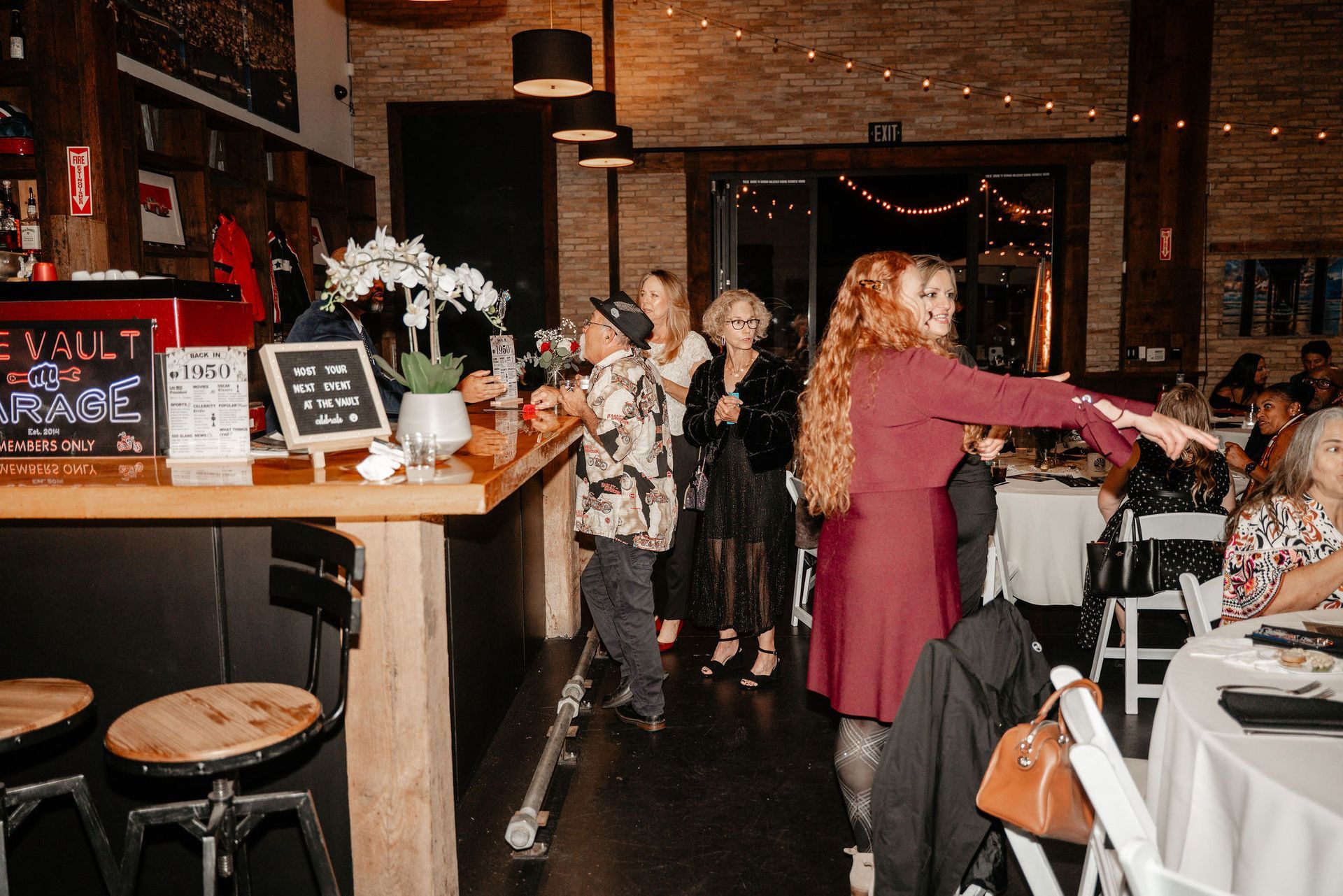 Interior event space; people at a bar and seated at tables. A woman in red gestures, others converse.