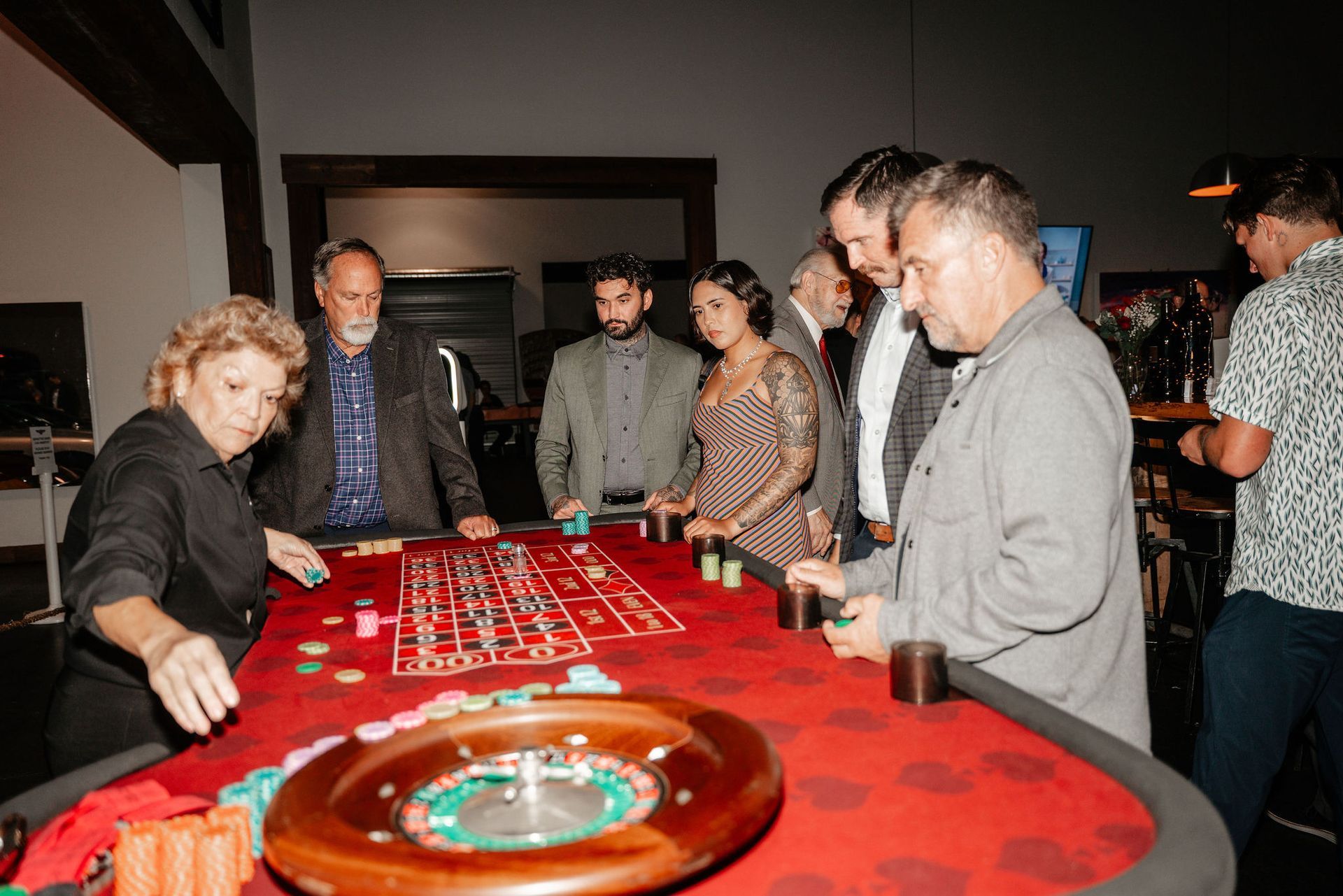 People gathered around a roulette table at a casino. Woman in black shirt dealing, others watching.