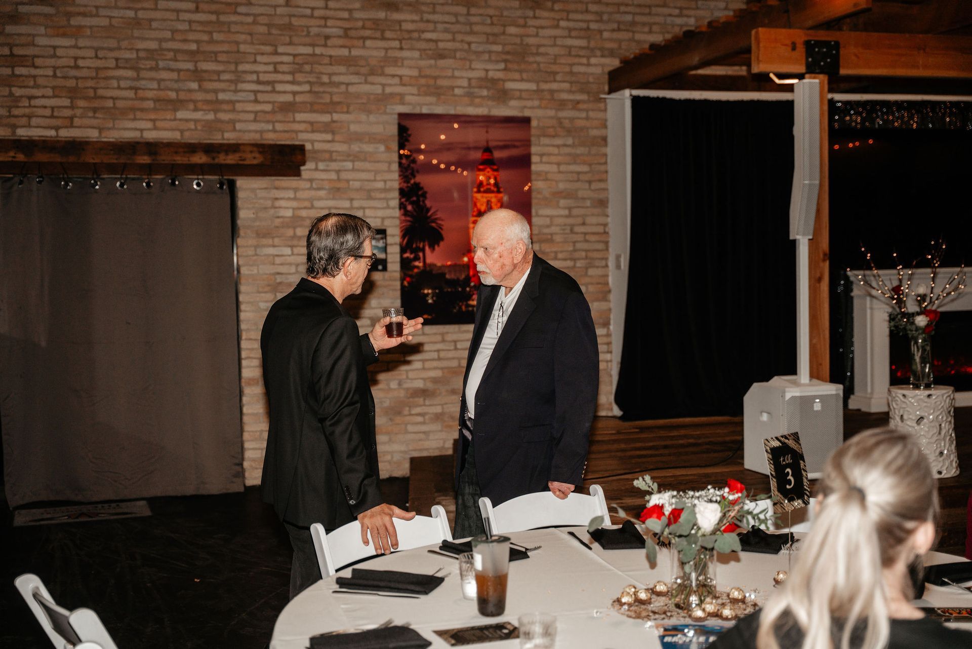 Two men in suits conversing at a table; a painting of Paris behind them.