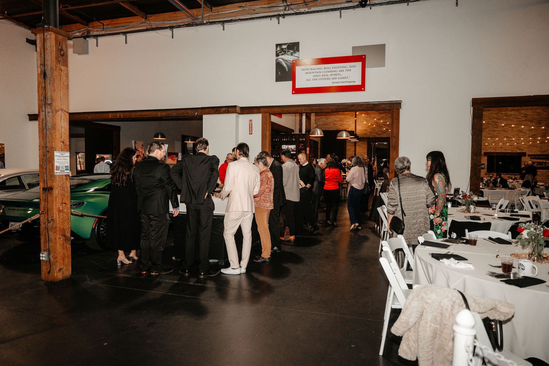 People gather at an event in a large room with tables, cars, and a doorway.
