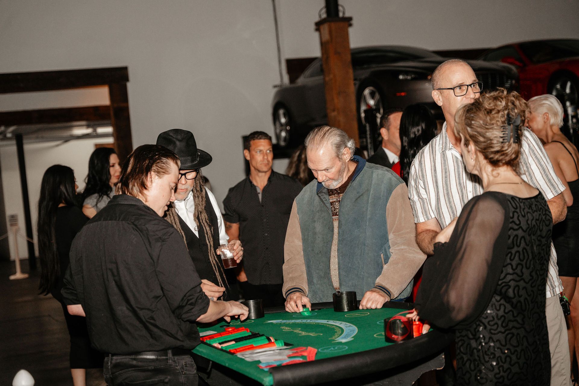 People gathered around a blackjack table in a room. Cars in background.