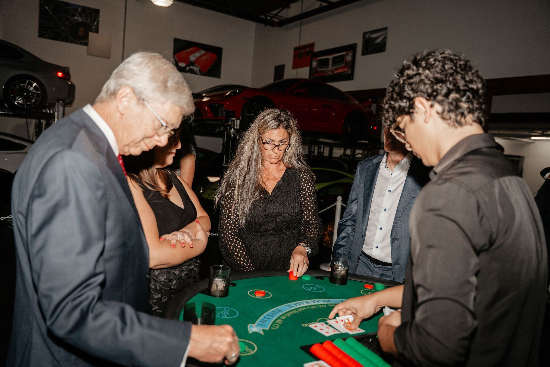 People playing blackjack at a party in a car garage; red and green chips visible on the table.