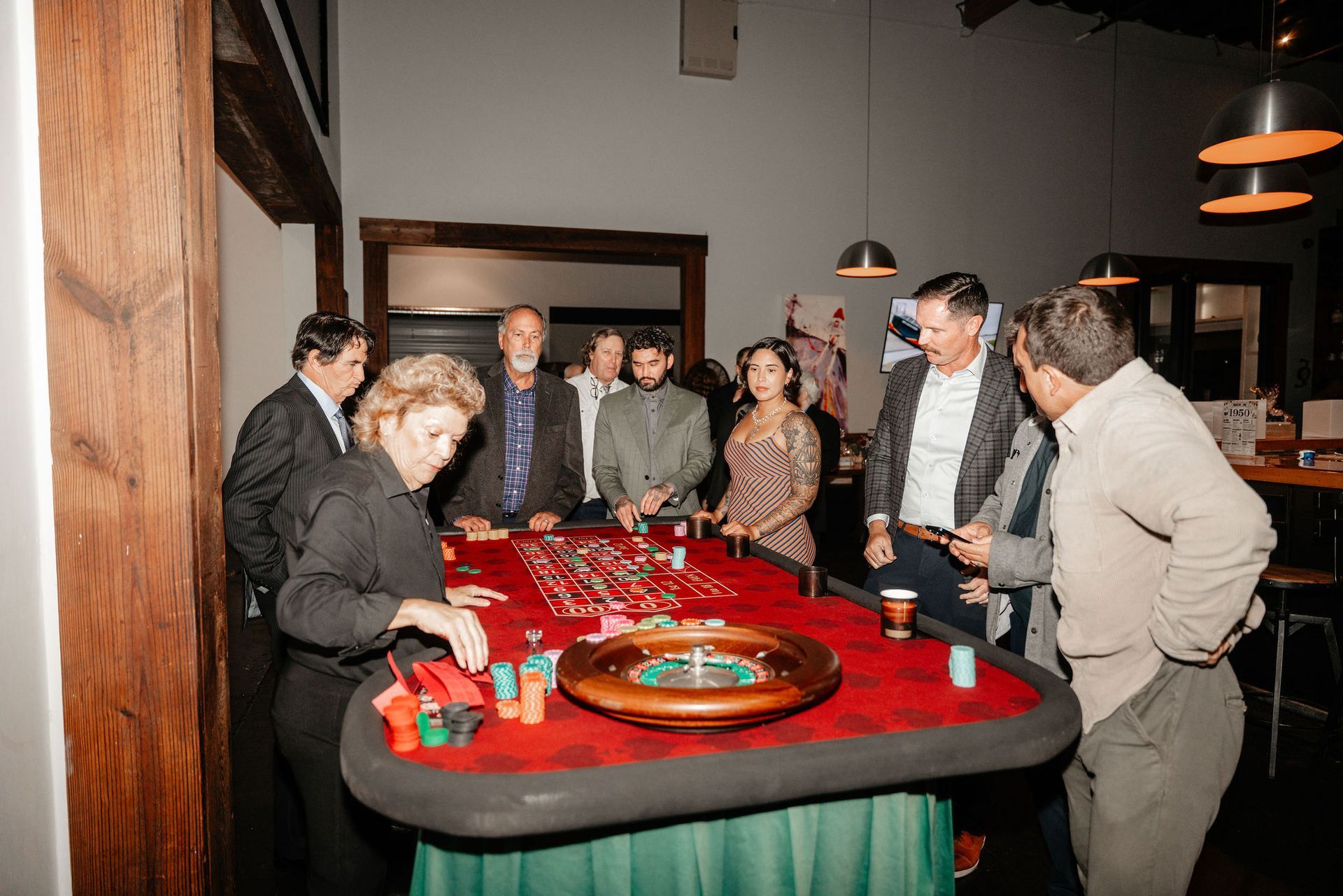 People gathered around a roulette table with a dealer; room has wood beams and pendant lights.
