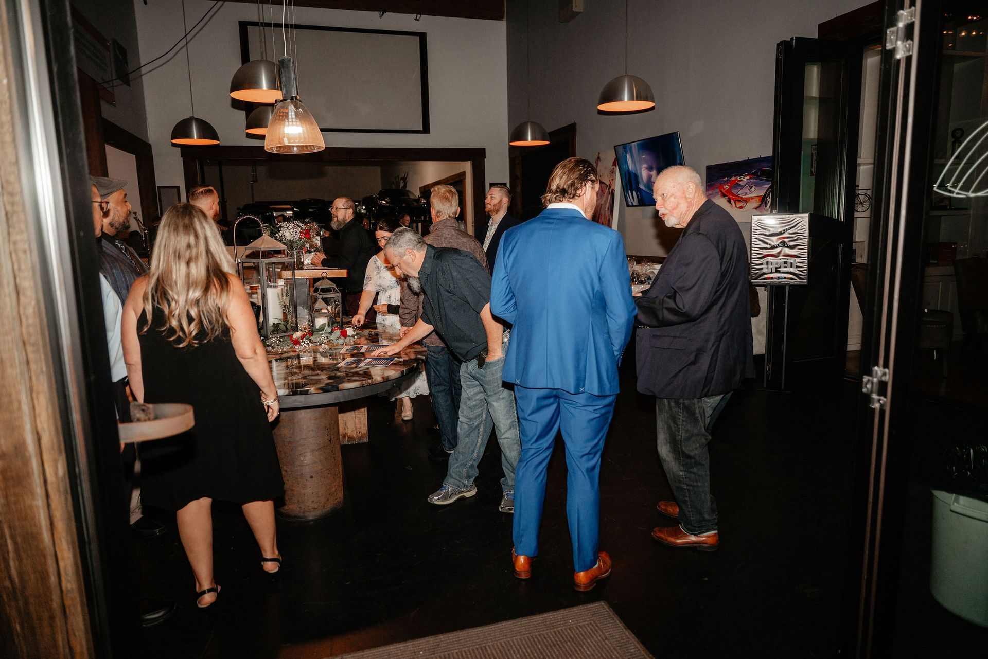 People gathered at an indoor event, near a table with objects. Low lighting, some wearing suits.