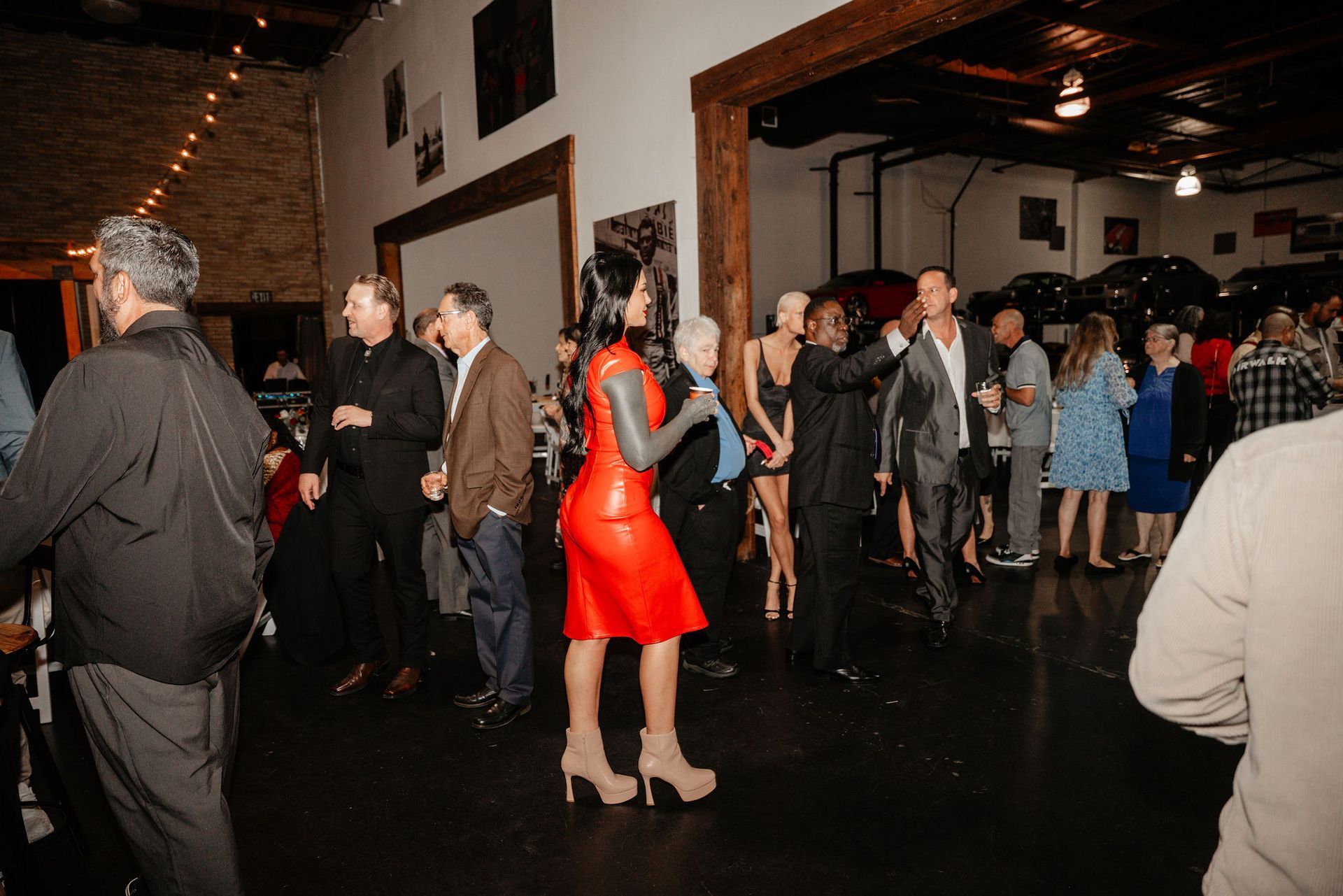 People at a social gathering in a room with vehicles. A woman in a red dress is in the foreground.