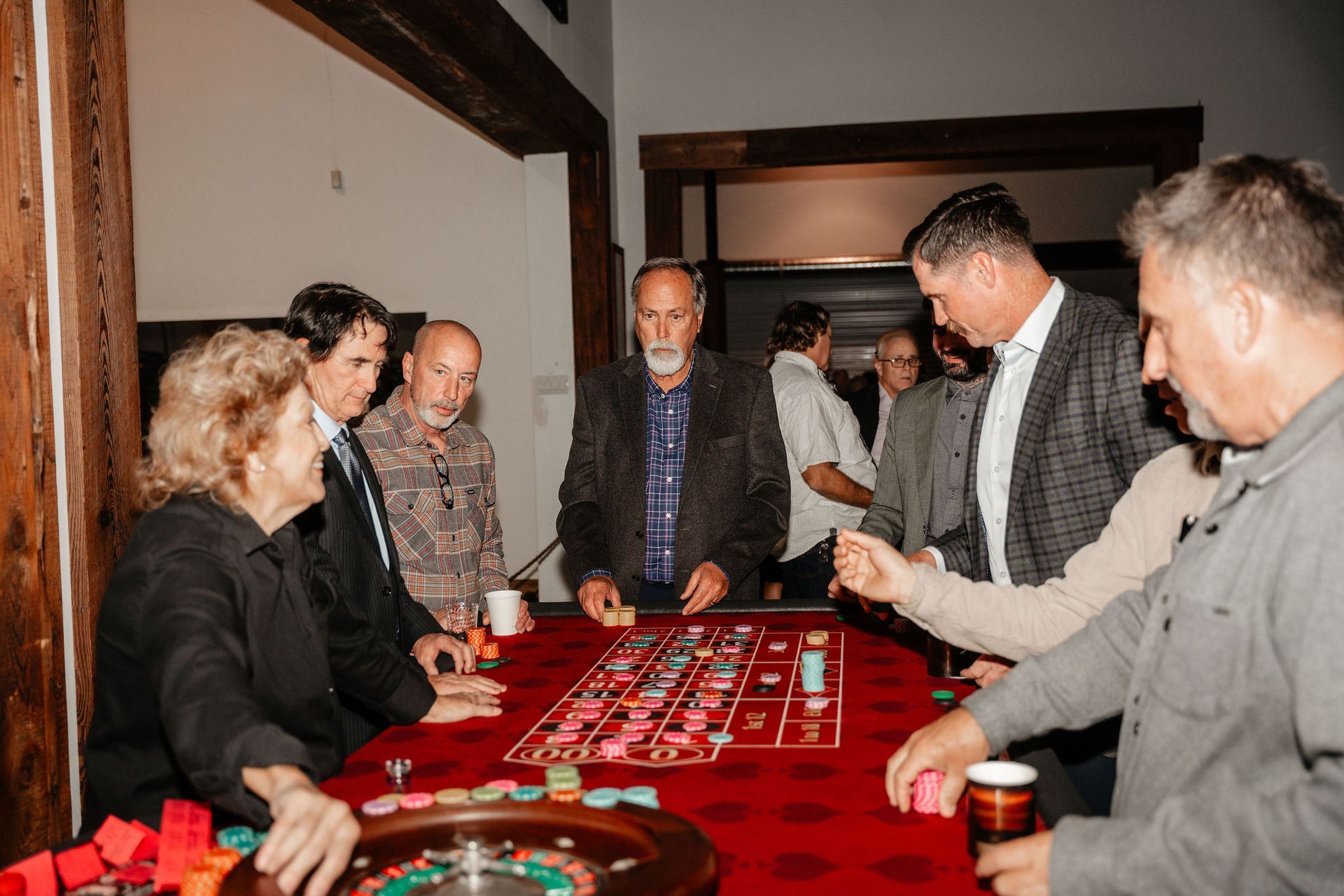 People gathered around a roulette table with chips in a room, some watching, some playing.