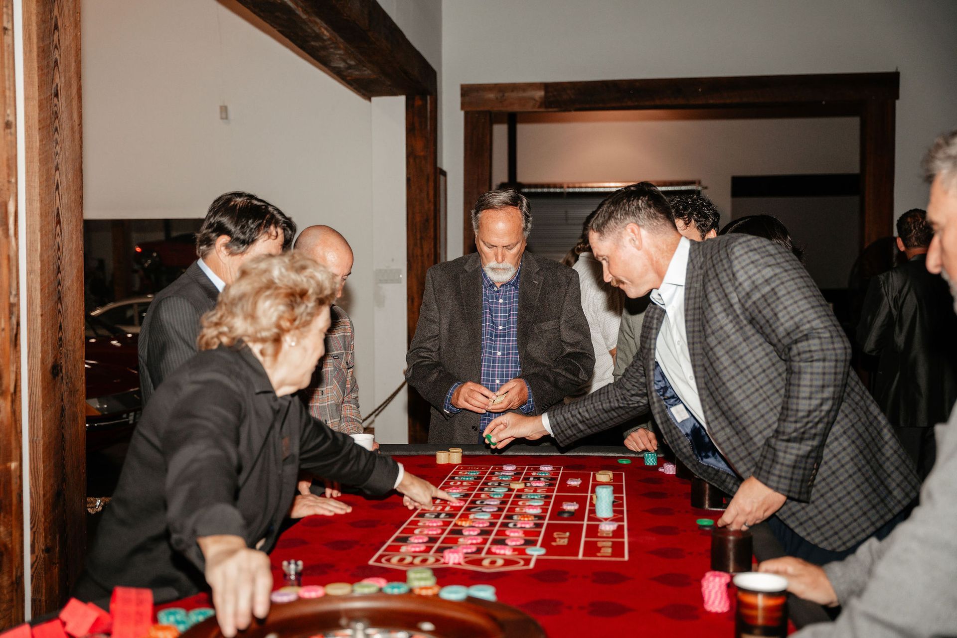 People playing roulette at a casino table. Red felt, colored chips, and players in suits. Interior with wooden beams.