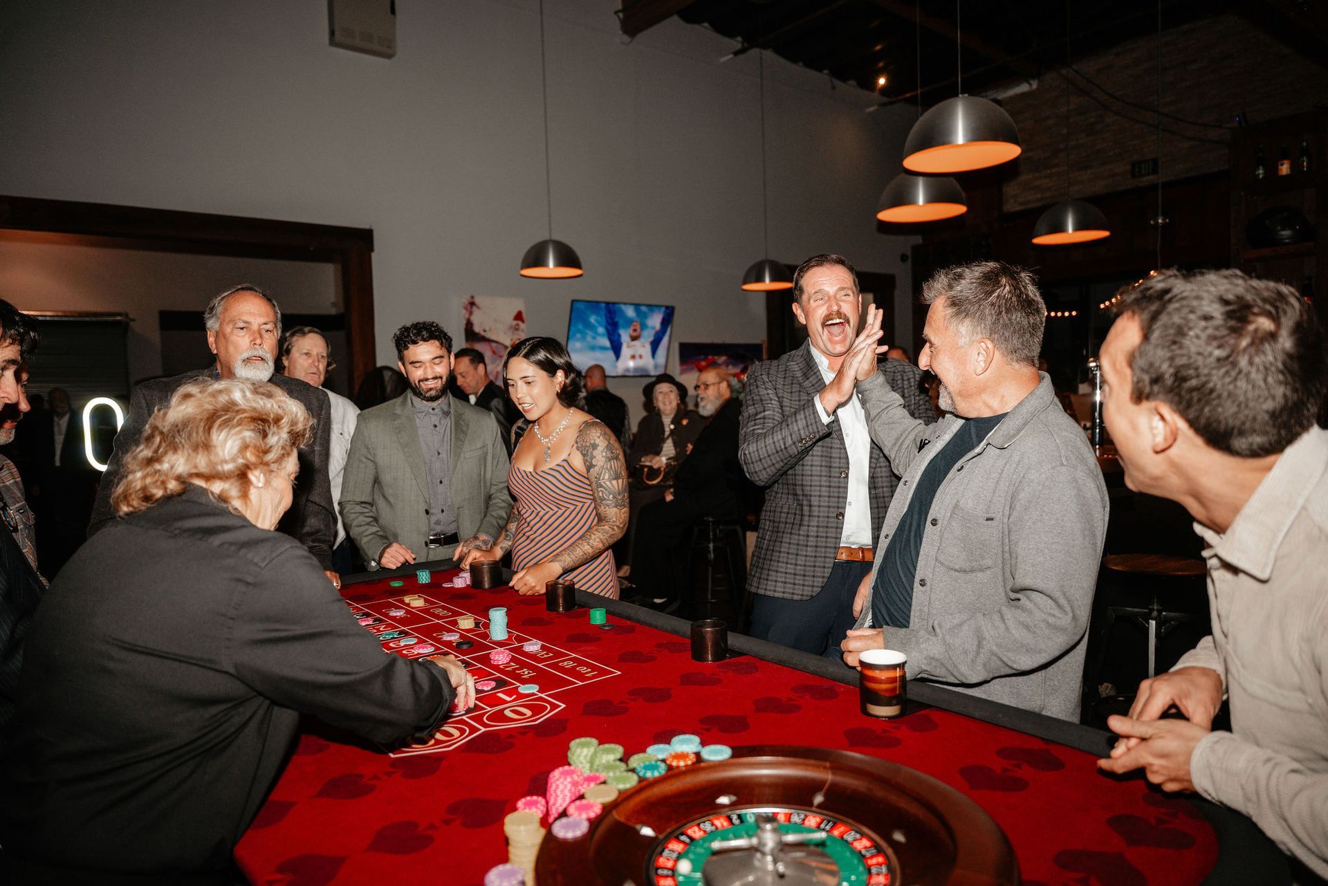 People at a roulette table in a dimly lit room, with one person clapping.